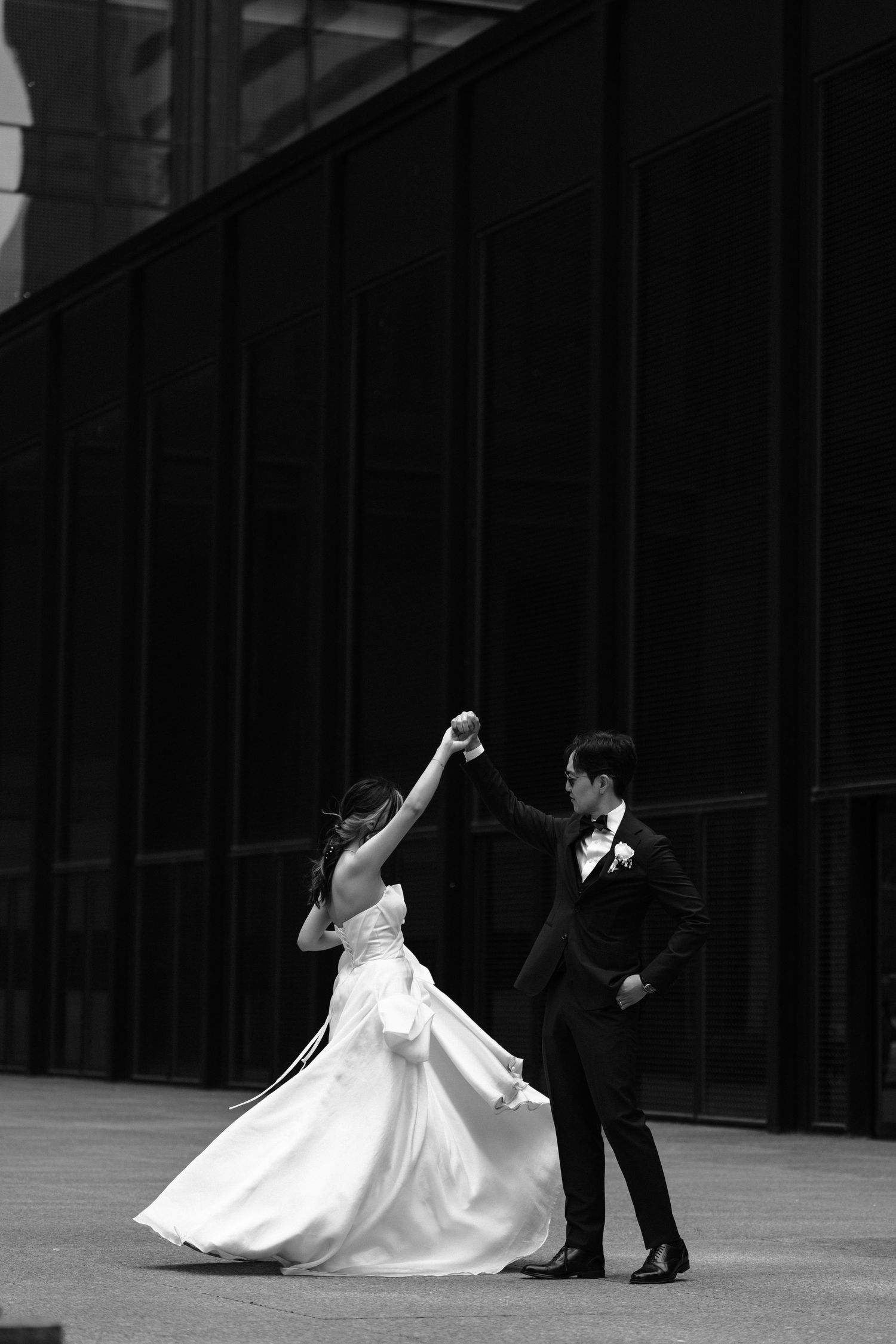A couple in wedding attire dances elegantly outside a modern building in black and white photography.