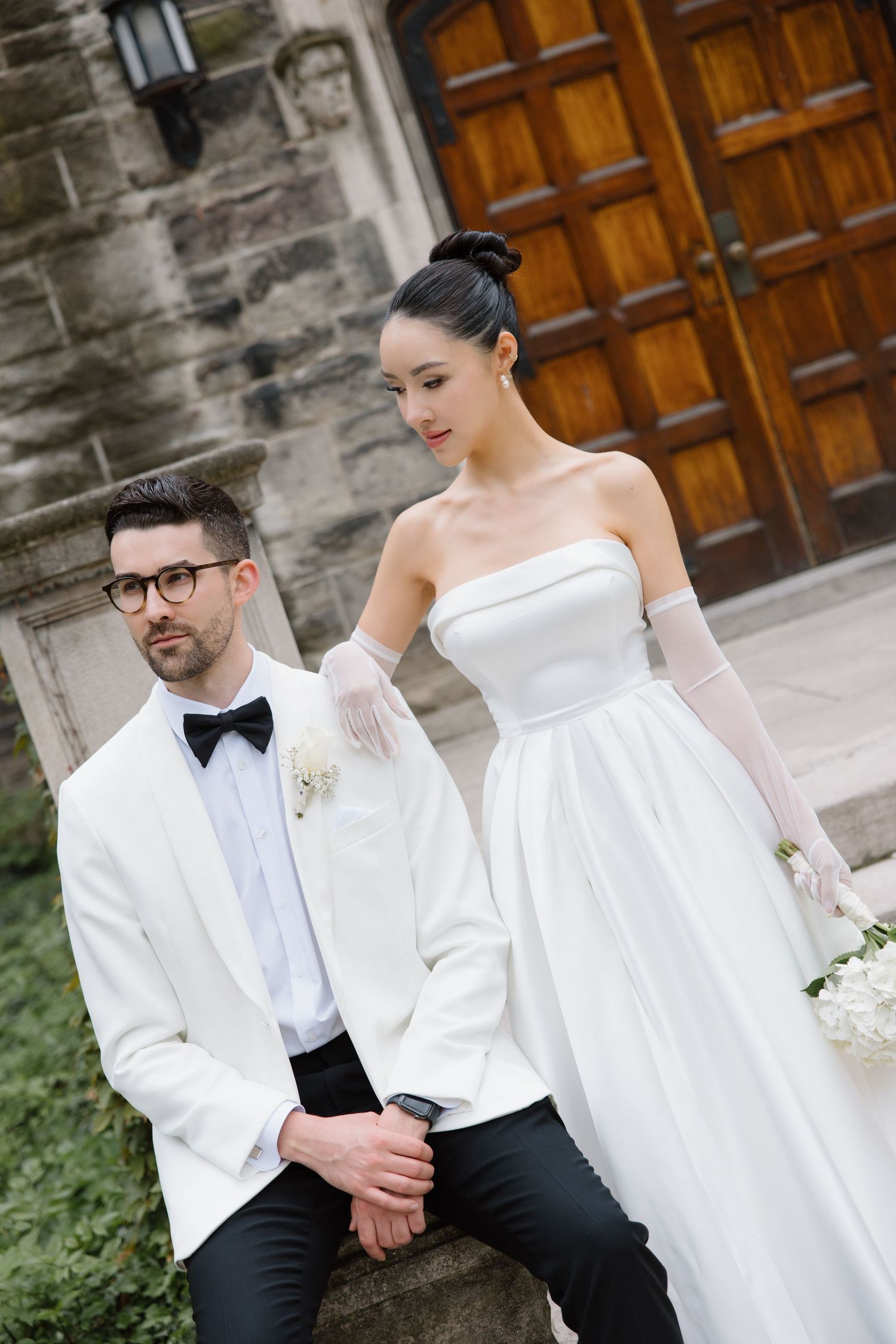 Elegant wedding couple in contrasting white and black attire pose outside historic stone building with wooden doors.