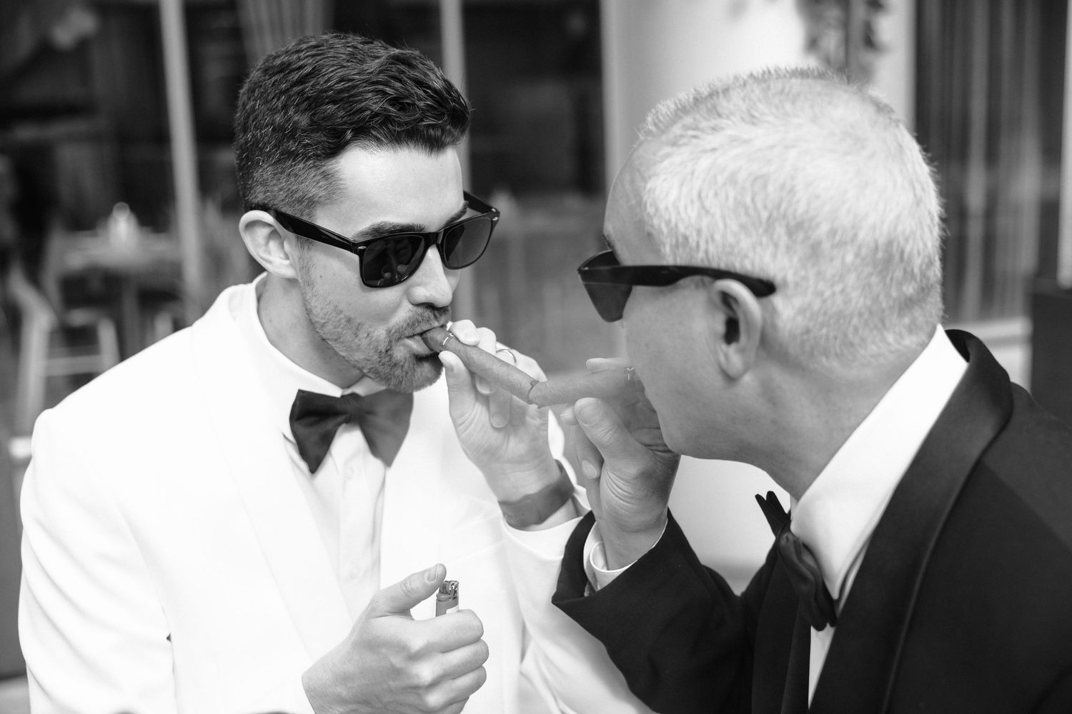 Two men in sunglasses and formal attire lighting cigarettes together in black and white vintage photograph.