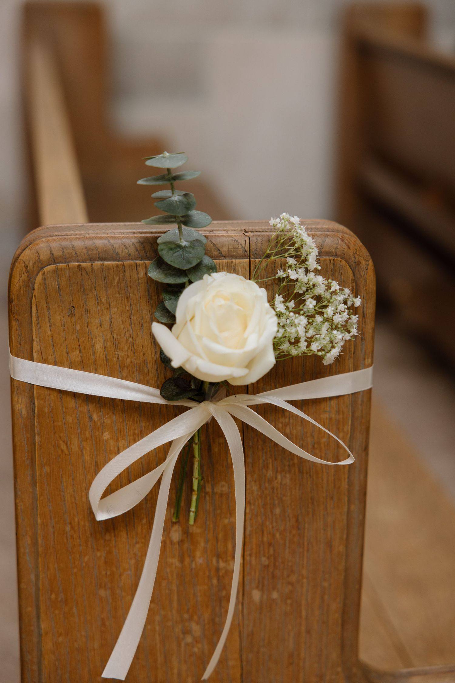 A white rose with eucalyptus and baby's breath tied with a cream ribbon on a wooden church pew for wedding decoration.