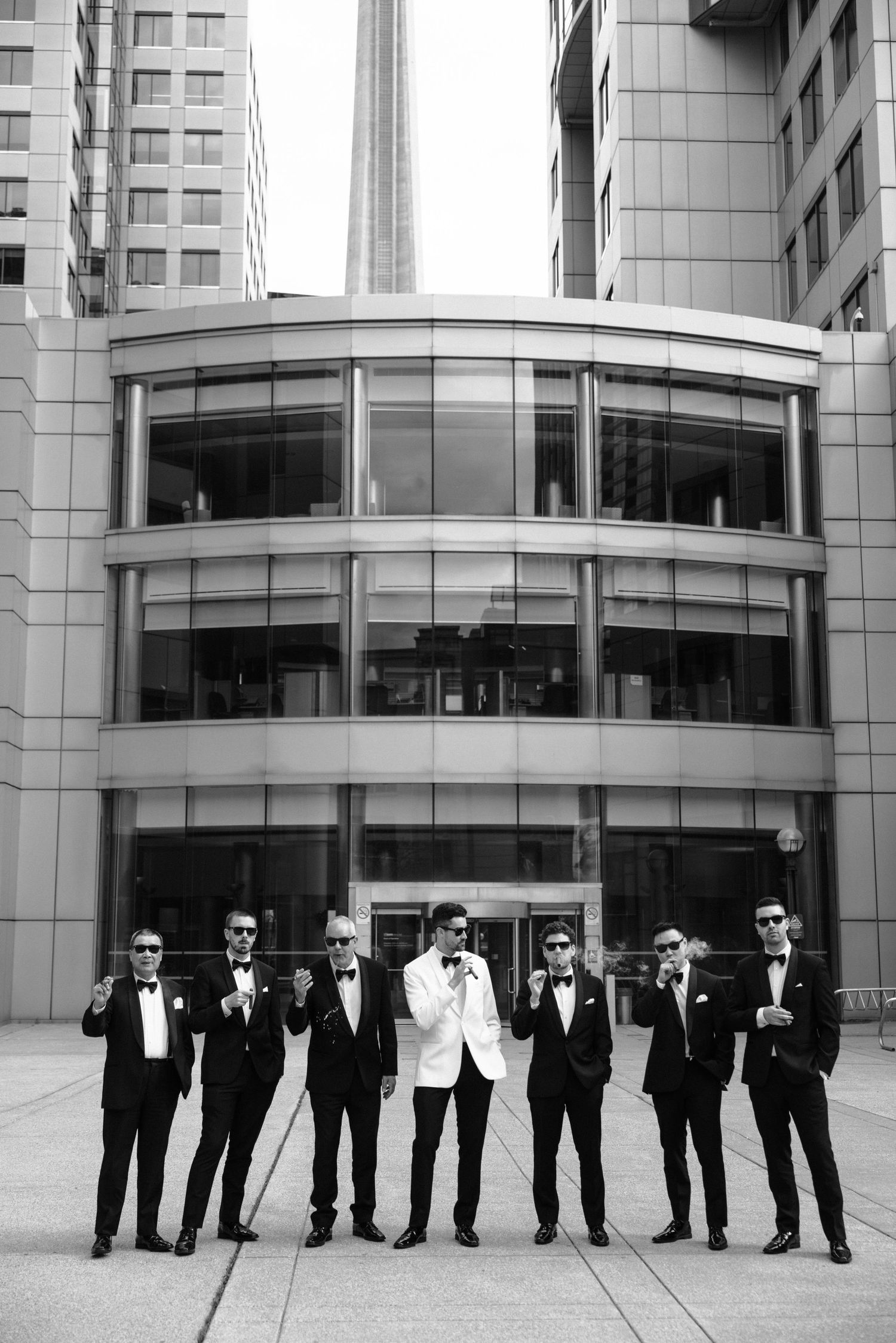 A row of groomsmen in black suits and sunglasses stand in front of a modern glass building exterior.