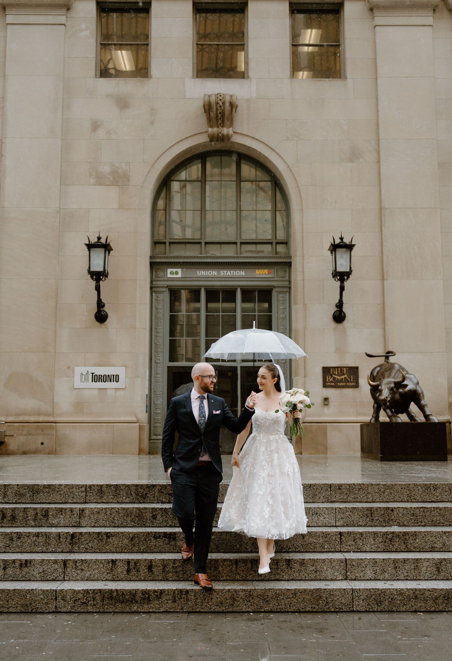 A couple stands on stone steps of a grand historic building, holding an umbrella in rainy weather.