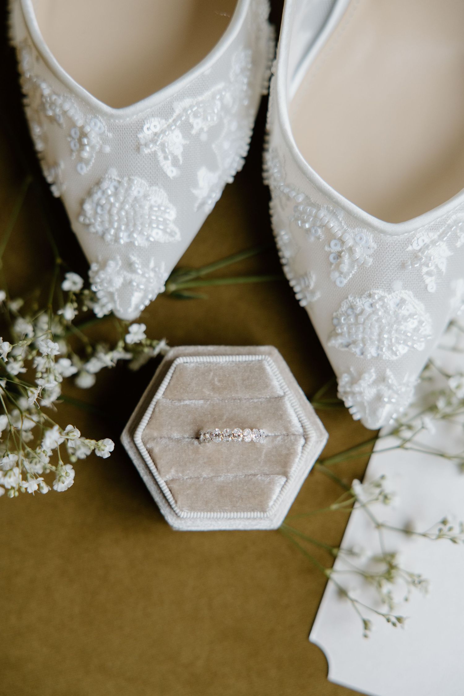 White lace bridal shoes with beaded details displayed alongside delicate baby's breath flowers and a hexagonal ring box.
