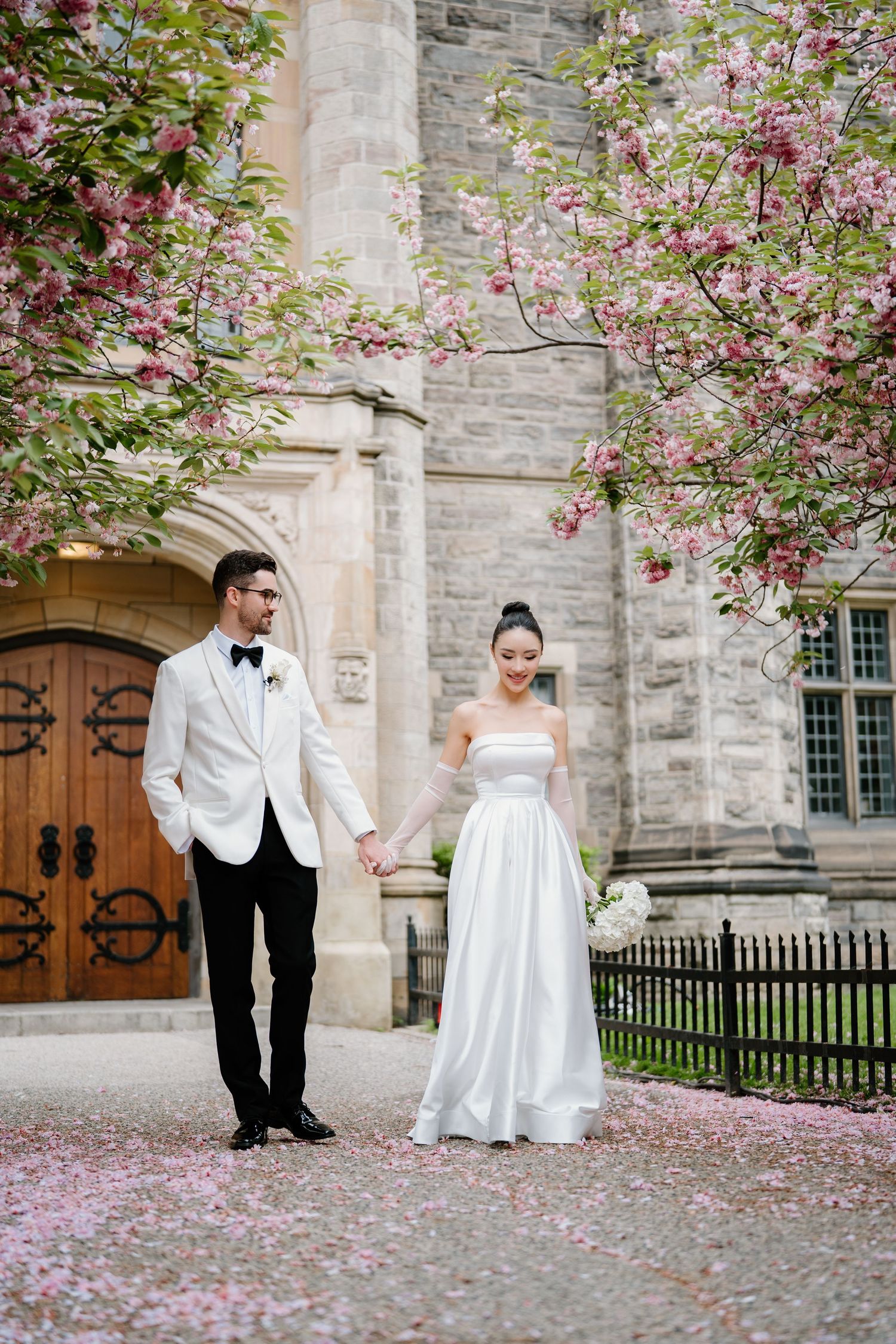 Wedding couple stands by a stone church wall with blooming pink cherry blossoms creating a romantic spring scene.