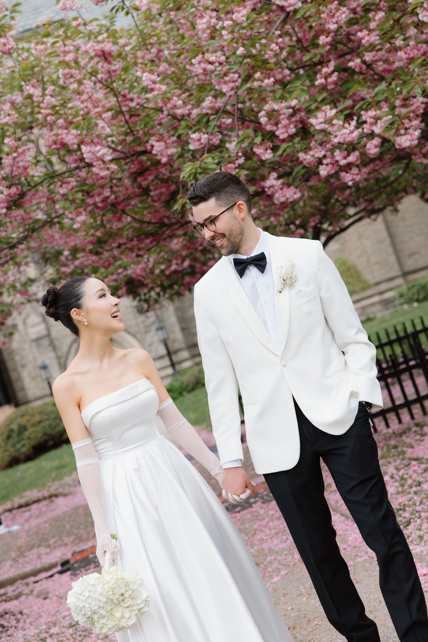 A wedding couple stands beneath blooming cherry blossoms on their special day, surrounded by pink petals.