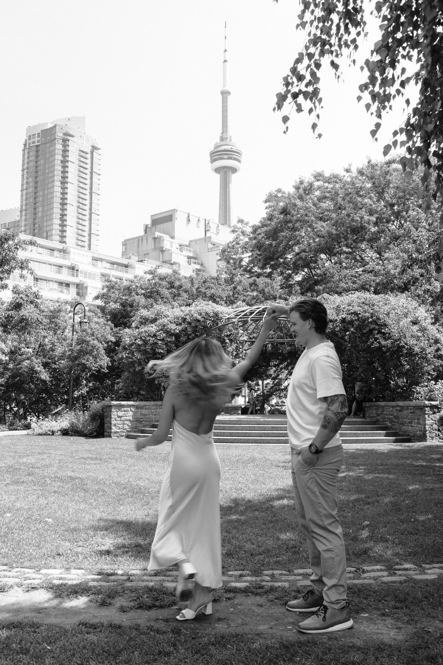 A couple shares a playful moment in front of the CN Tower and Toronto skyline on a sunny summer day.
