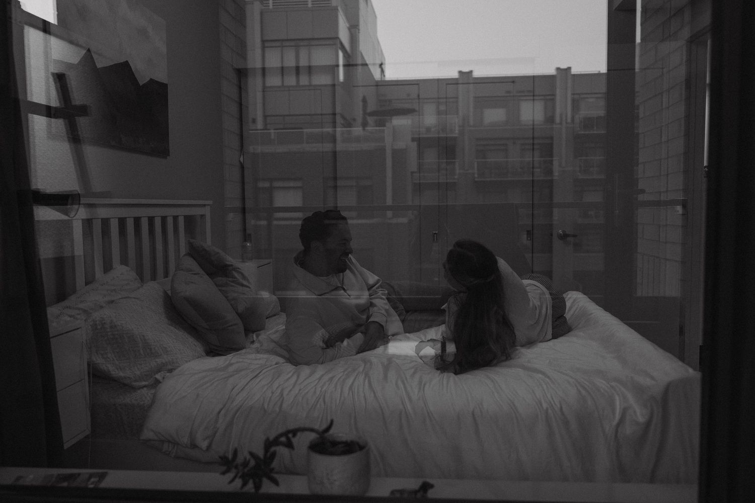 Two people sit together on a bed in a moody black and white urban apartment setting with city views through windows.