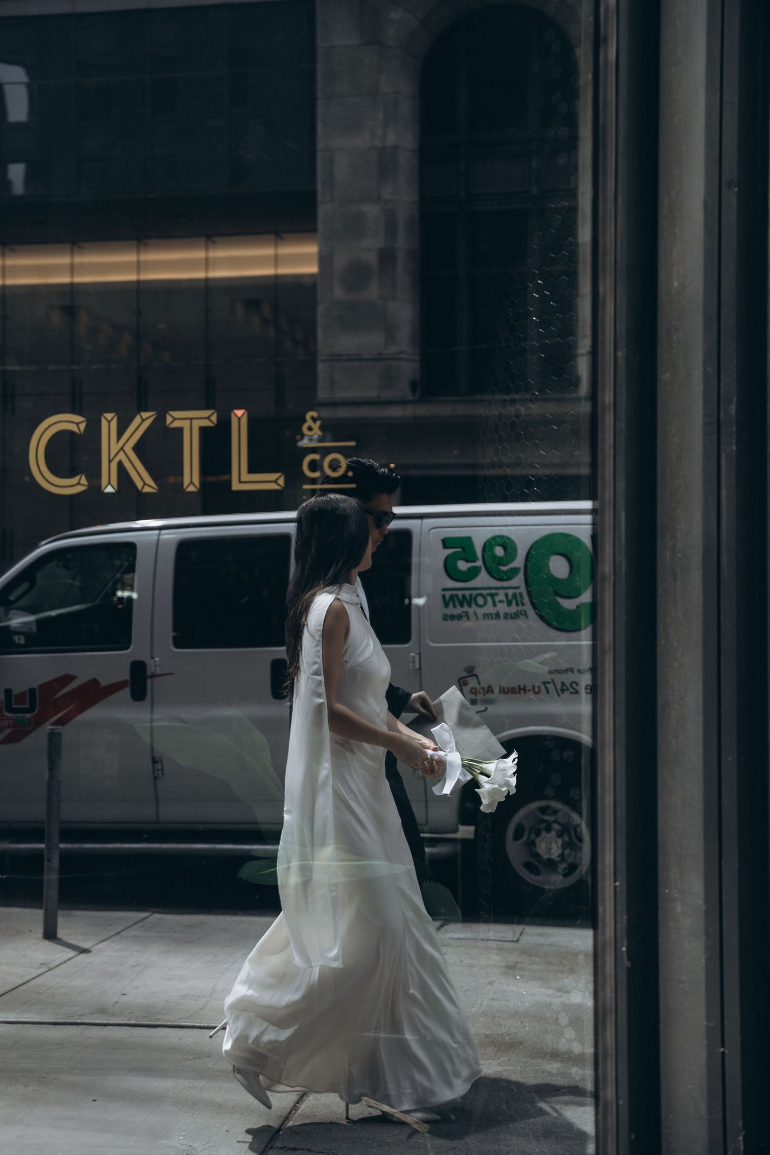 A person in a flowing white dress walks past an urban storefront window with CKTL signage and a parked van visible outside.