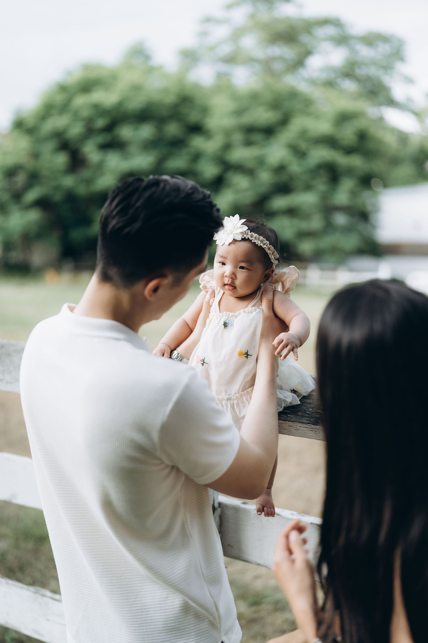 A baby in a white dress and floral headband gets lifted up near a wooden fence on a sunny day.