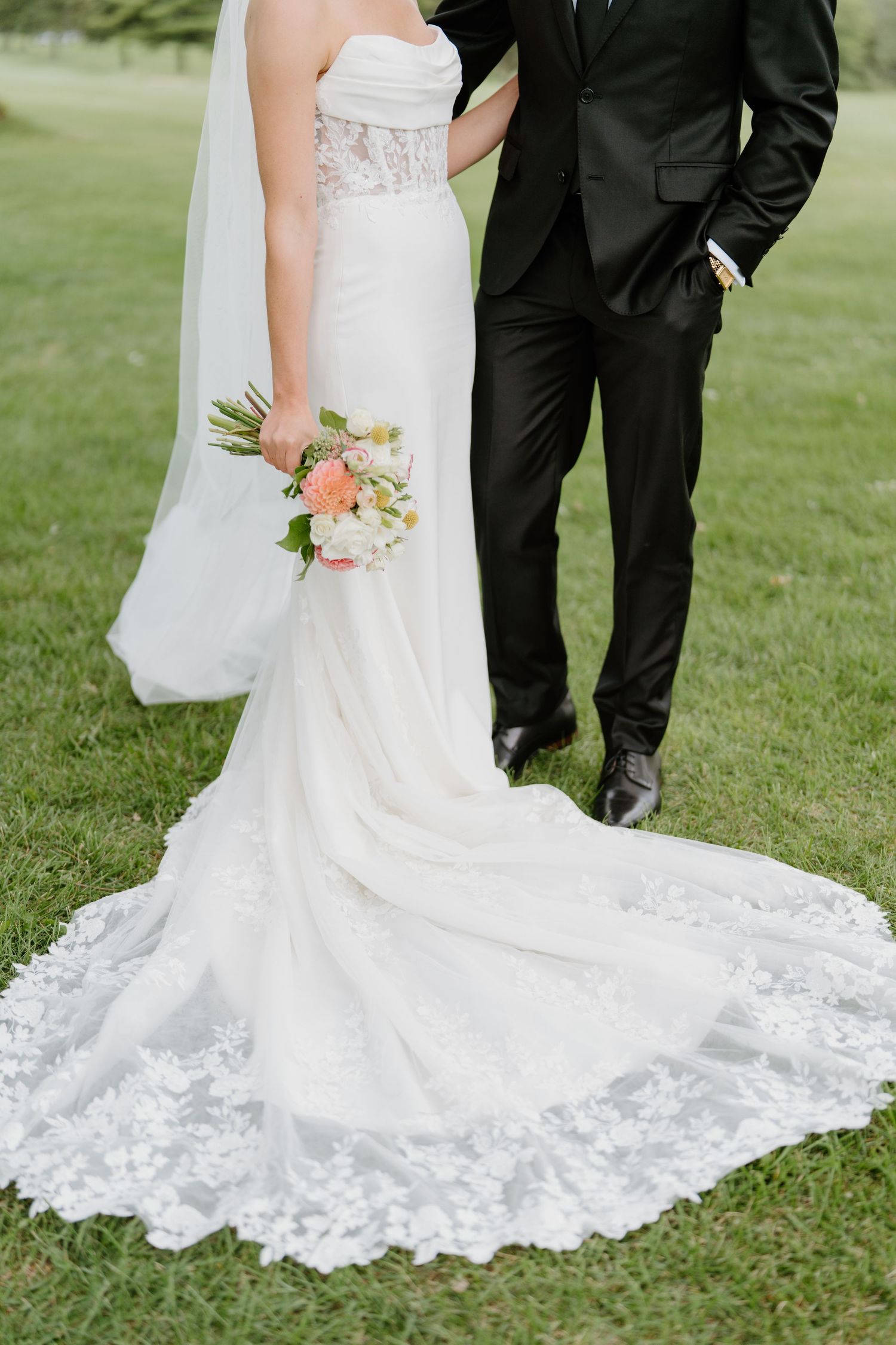 Elegant white lace wedding dress with a dramatic train spreads across green grass beside black formal attire.