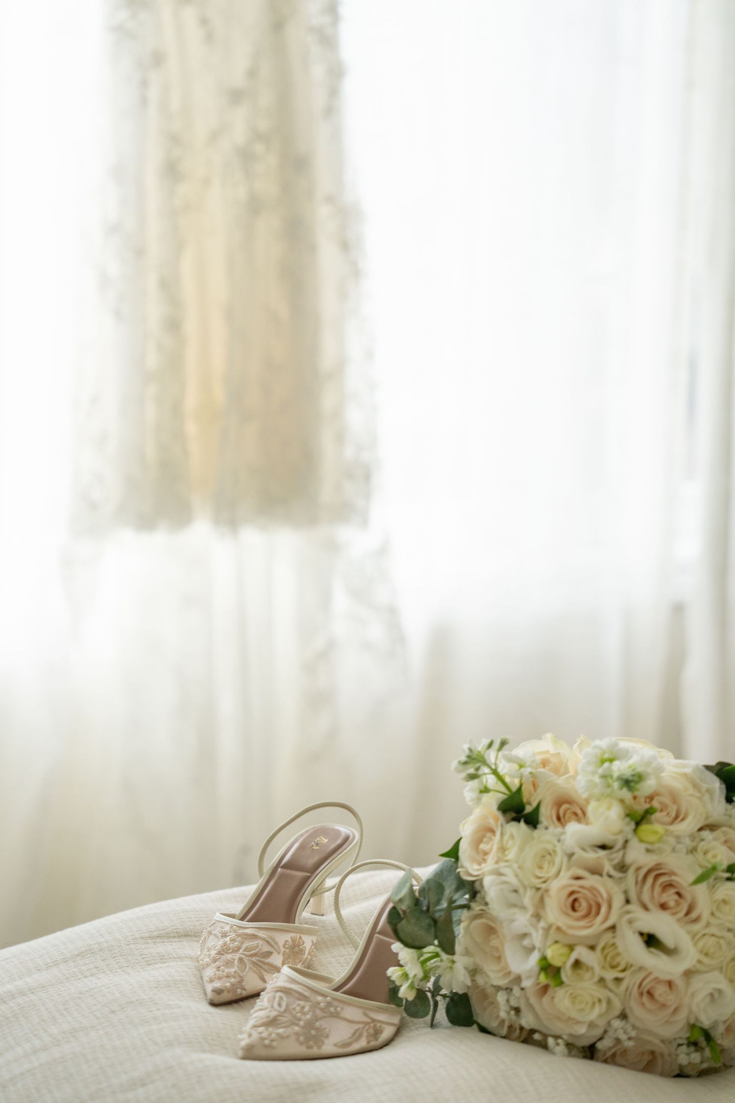 Elegant bridal shoes and white rose bouquet arranged on white bedding with soft window light in background.