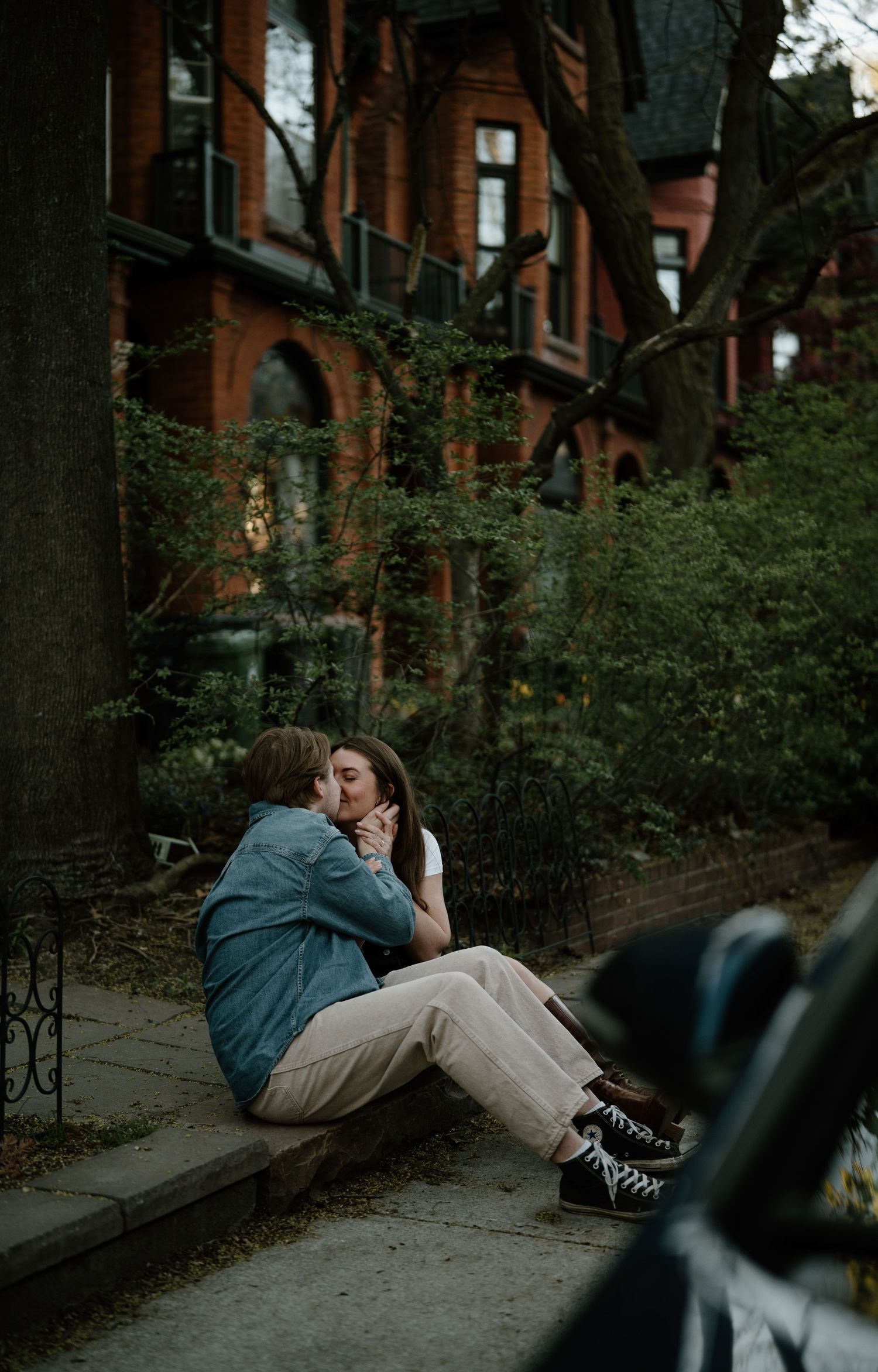 Two people embrace on a sidewalk near ivy-covered brick buildings at dusk.