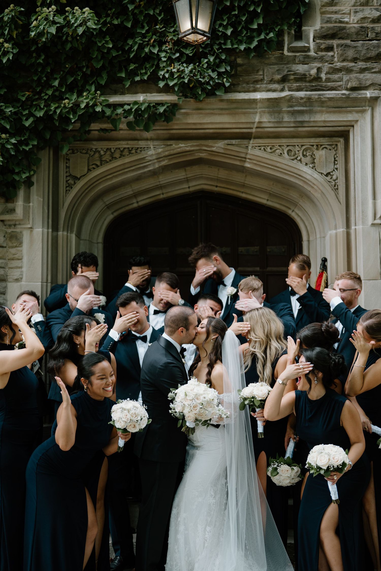 Wedding party in formal black attire and white bridal gown pose together outside stone building with ornate doorway.