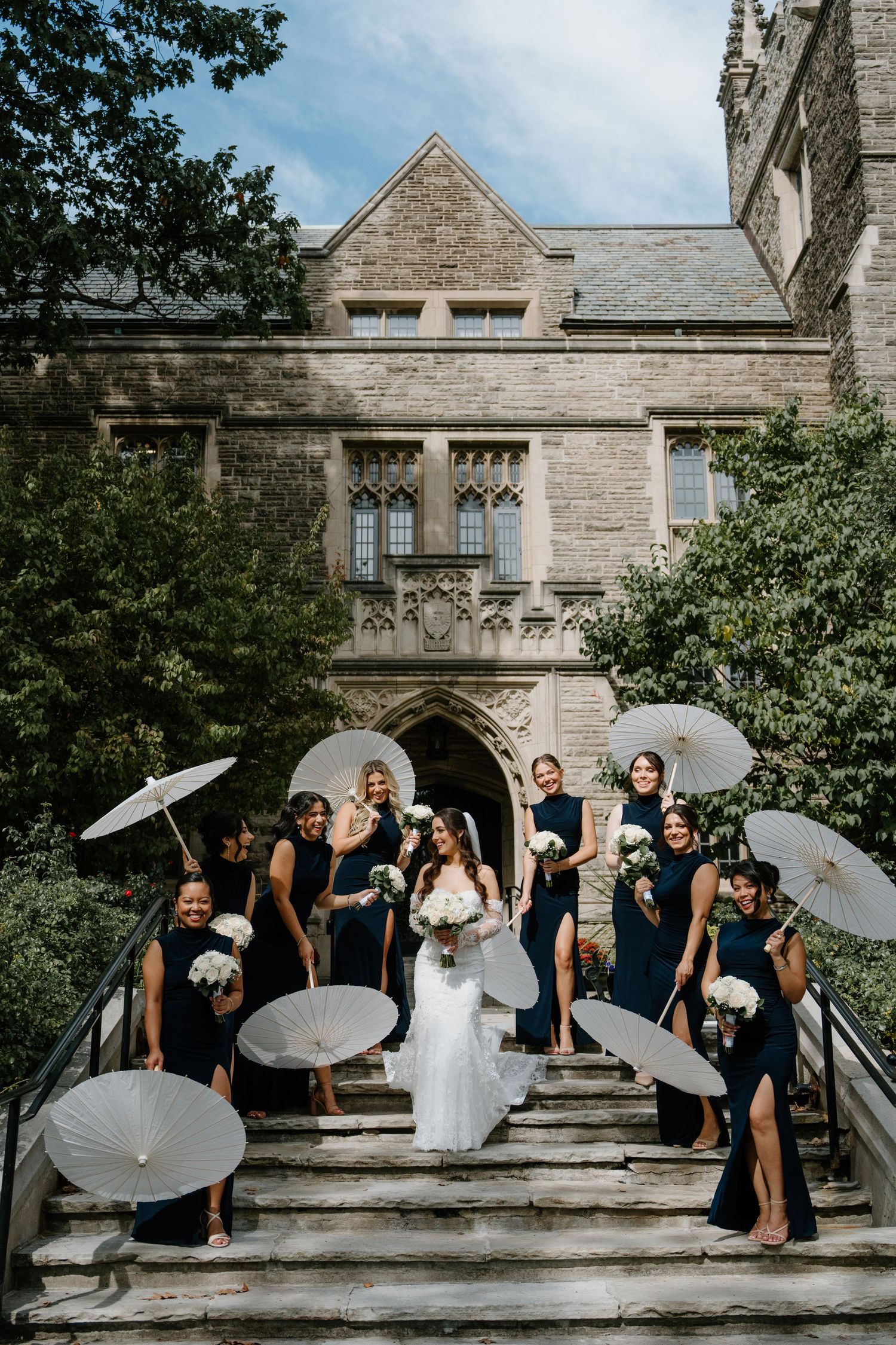 Wedding party poses on stone steps in sequence with white parasols against Gothic architecture backdrop.