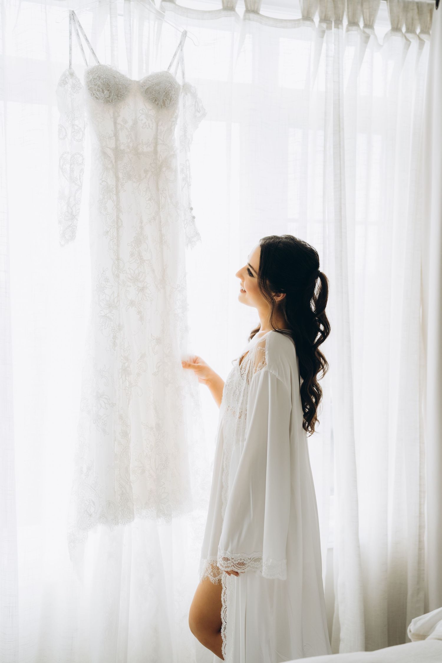 Figure in flowing white robe examines elegant dresses hanging against sheer curtained window.