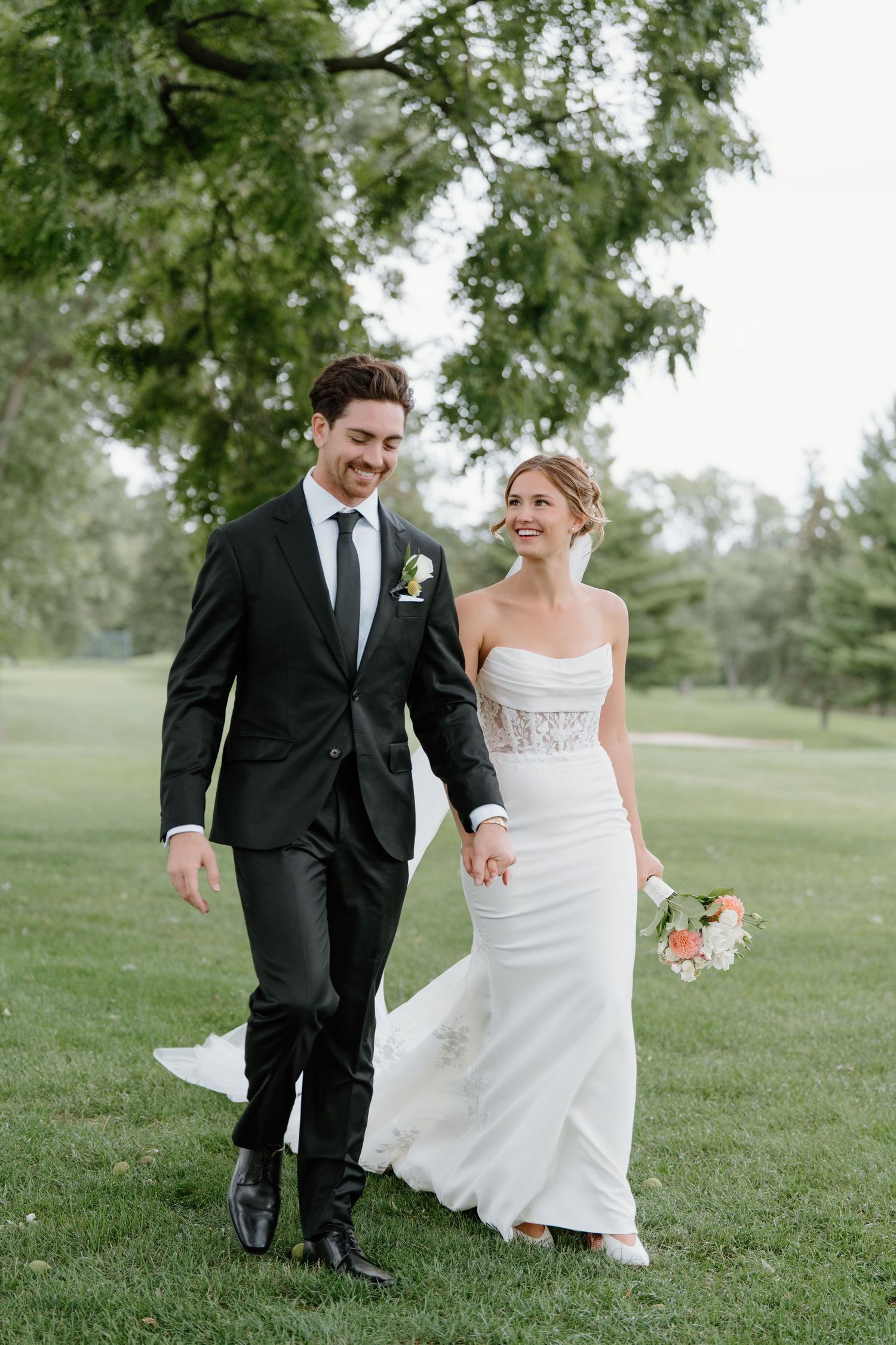A couple in wedding attire playfully walks and dances together on a grassy lawn surrounded by trees.