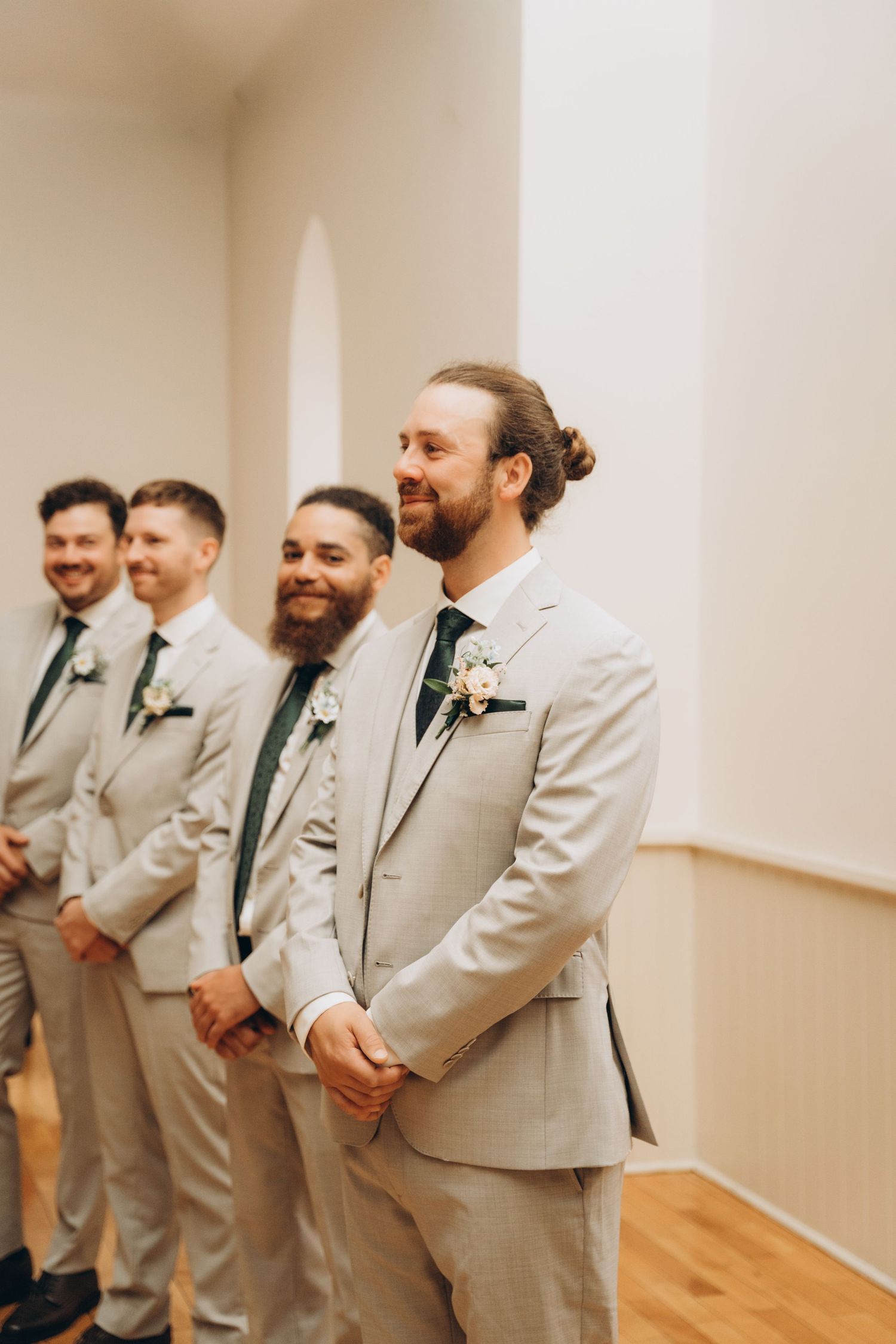 A line of groomsmen in light gray suits and dark ties stand together in a minimalist indoor setting.
