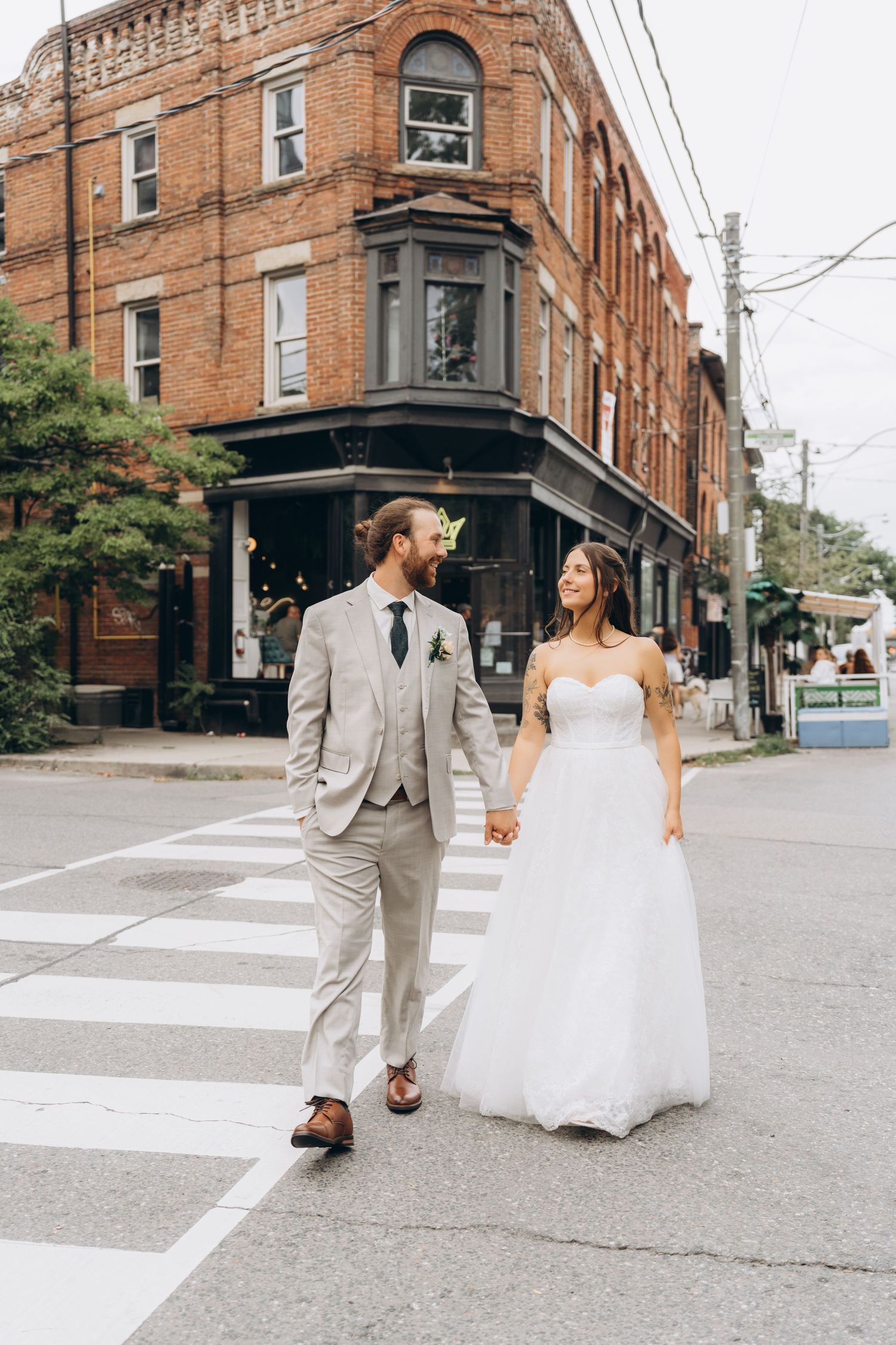 A couple in wedding attire walks hand in hand across a crosswalk by a historic brick building on a city corner.