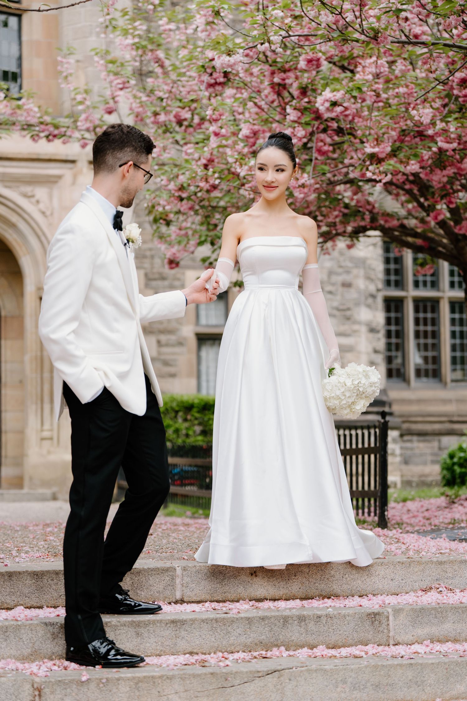 A couple in wedding attire stands on stone steps with blooming pink cherry blossoms against a stone building.