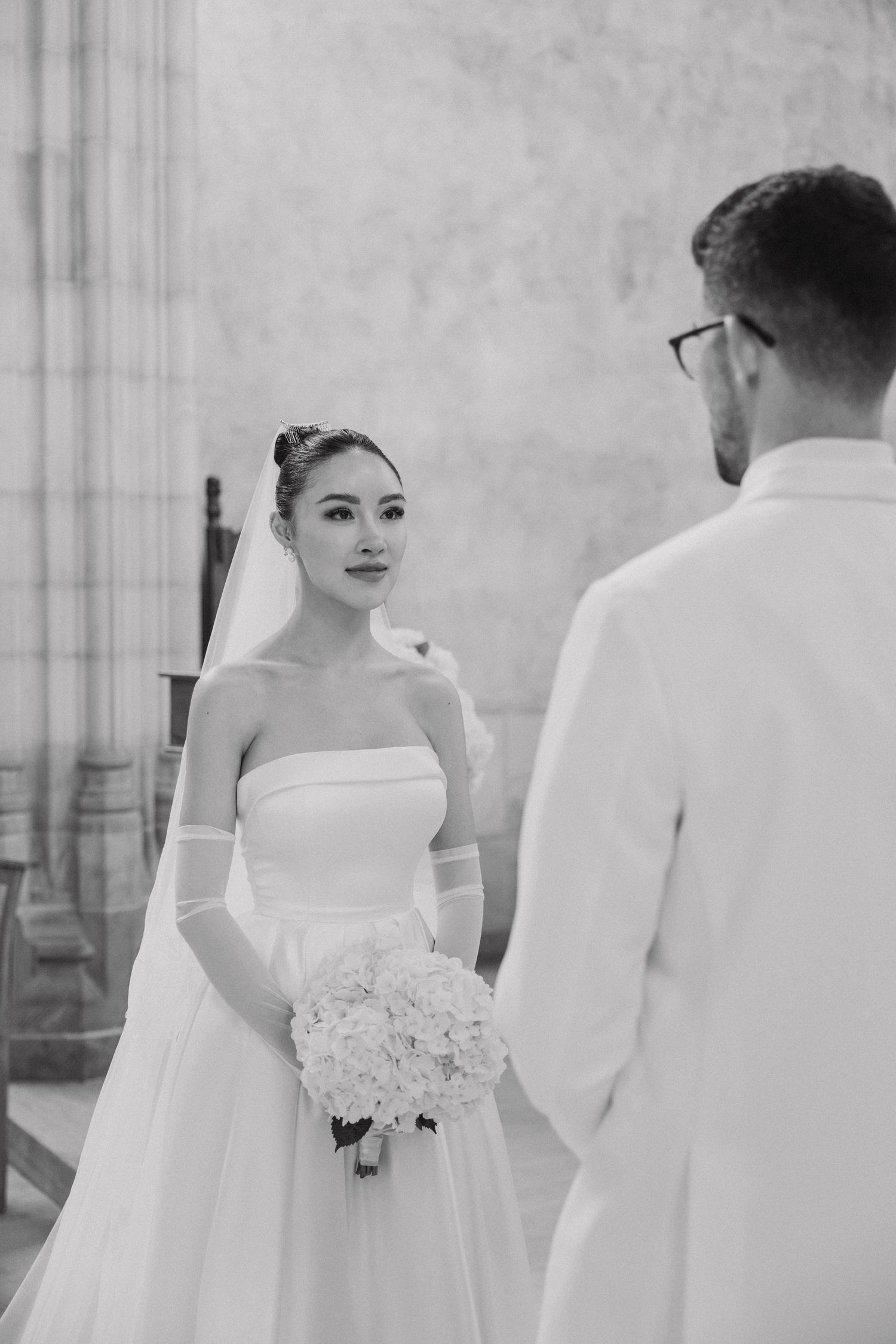A black and white wedding photo showing a bride in a strapless gown holding a bouquet facing her groom in a white suit.