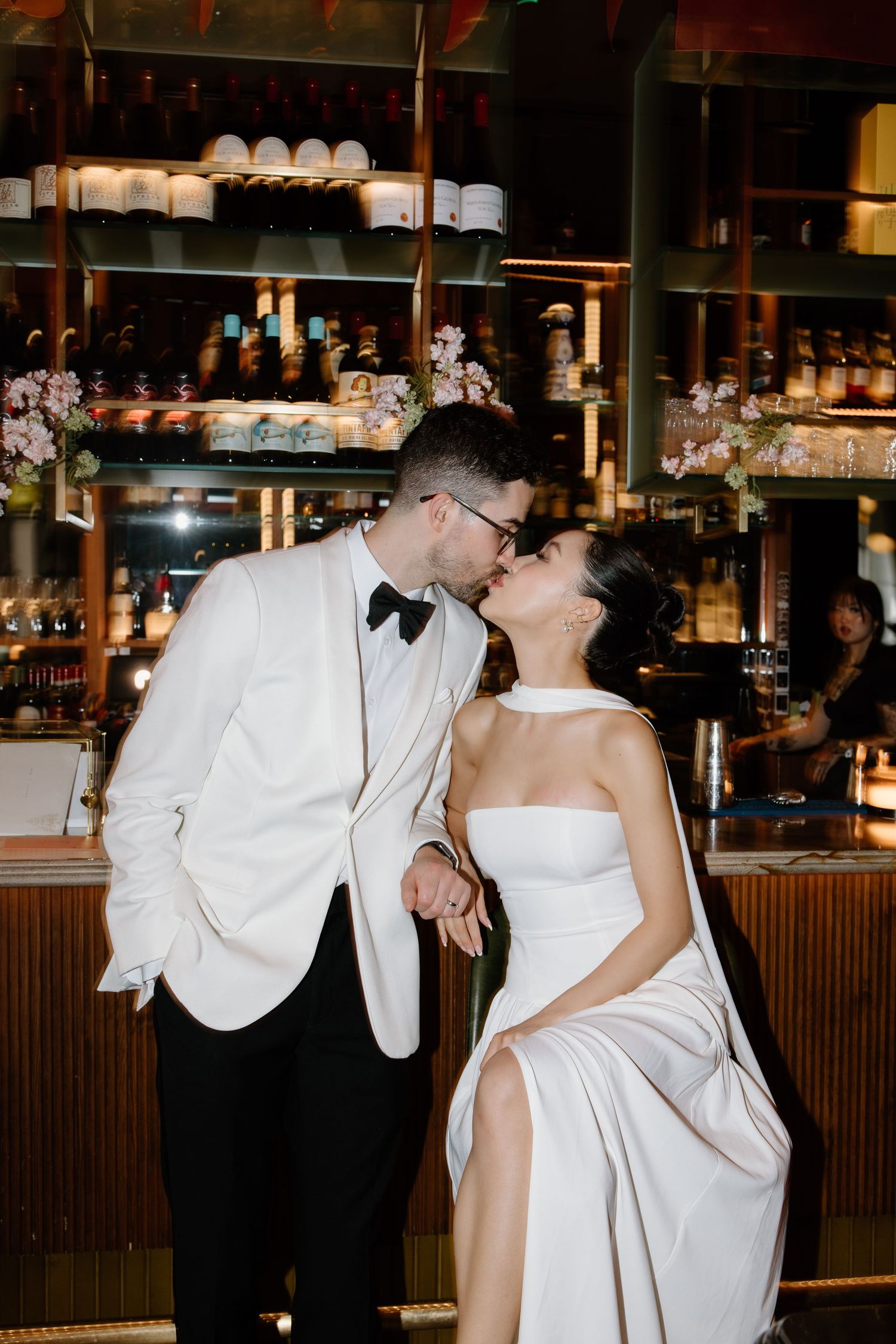 Romantic couple sharing an intimate moment at a bar with shelves of glassware and bottles in the background.
