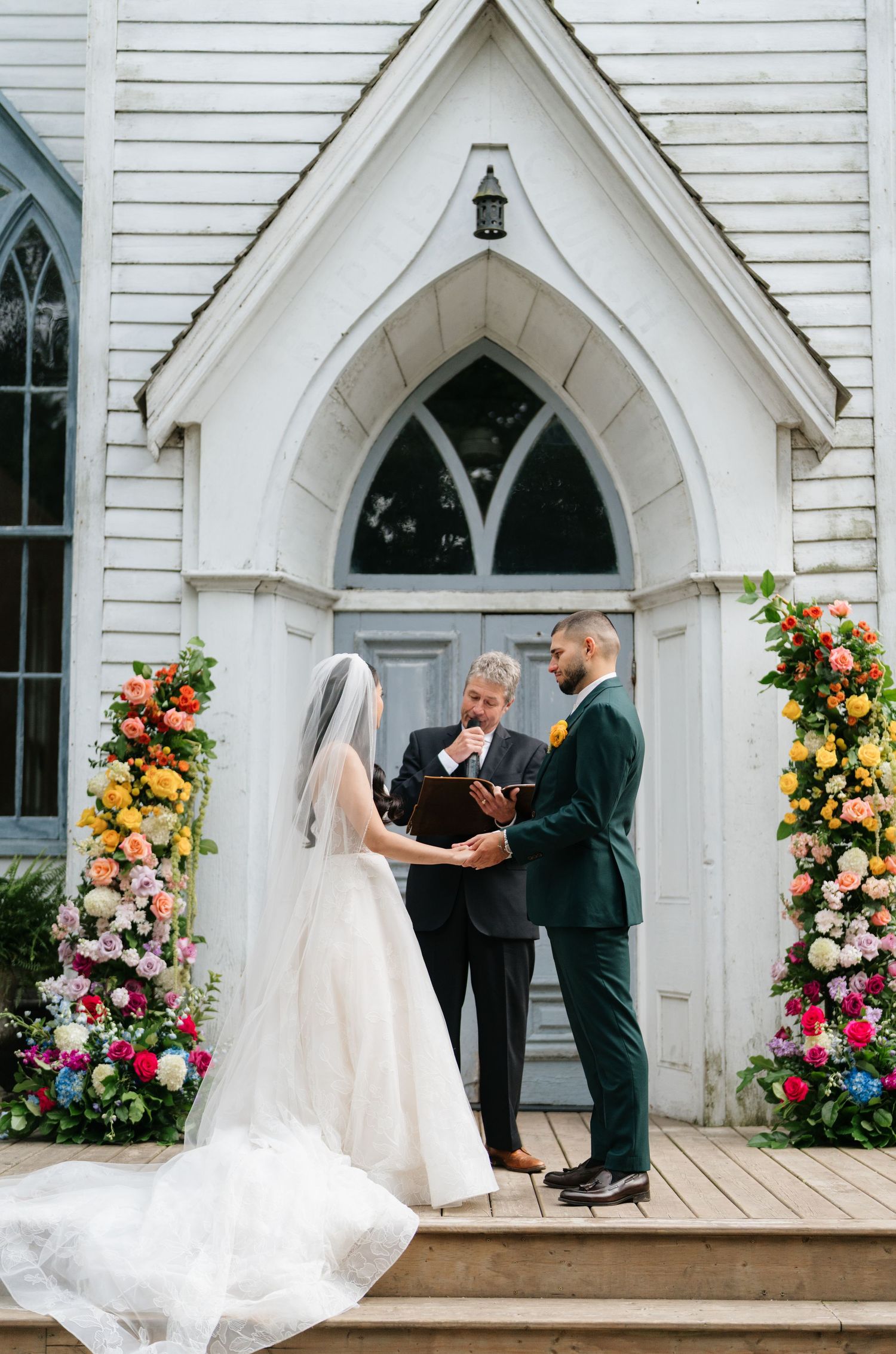 A romantic wedding ceremony takes place at a charming white chapel with Gothic arched windows and colorful floral arrangements.