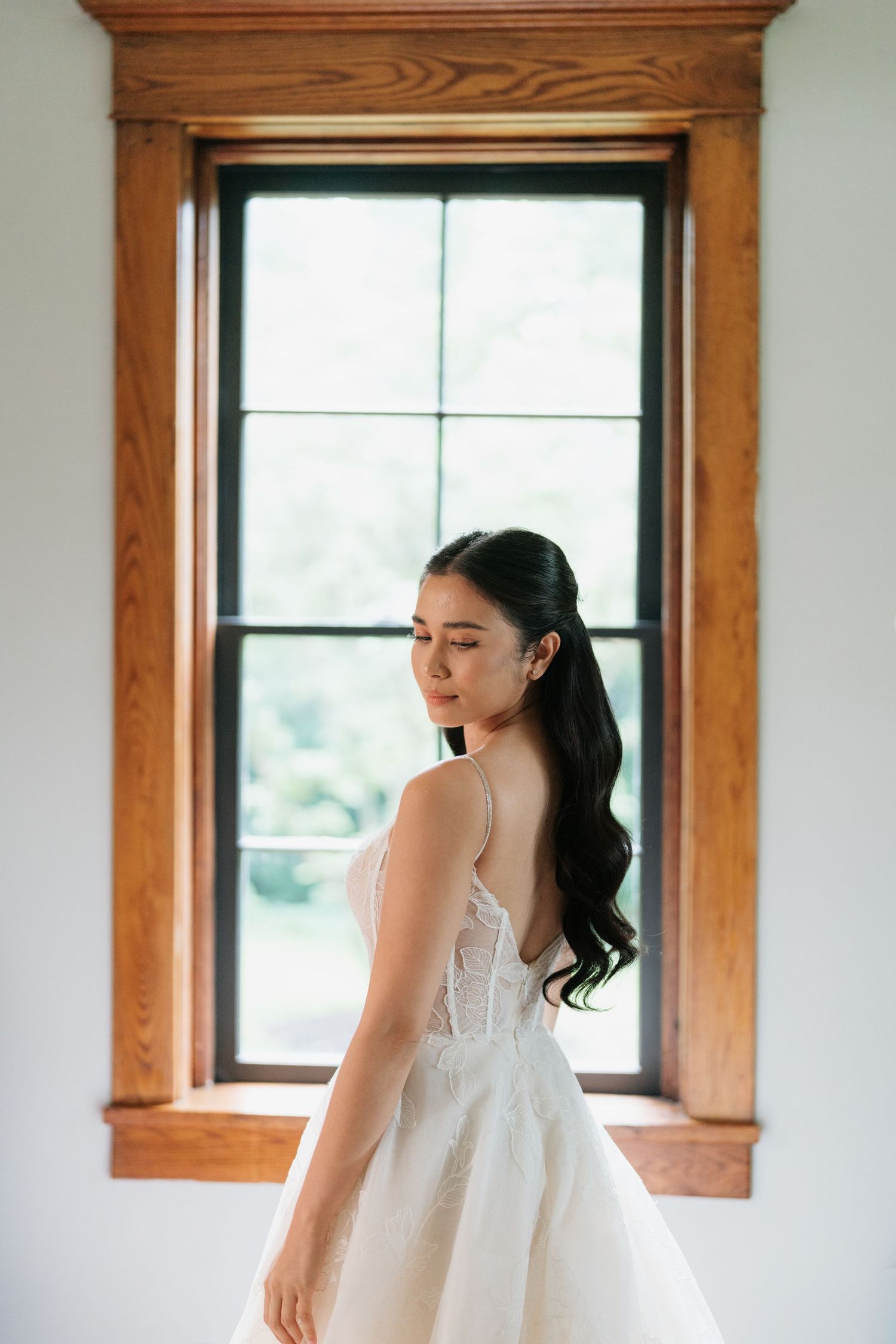 A bride in a white wedding dress stands elegantly near wooden-framed windows in a bright, sunlit room.
