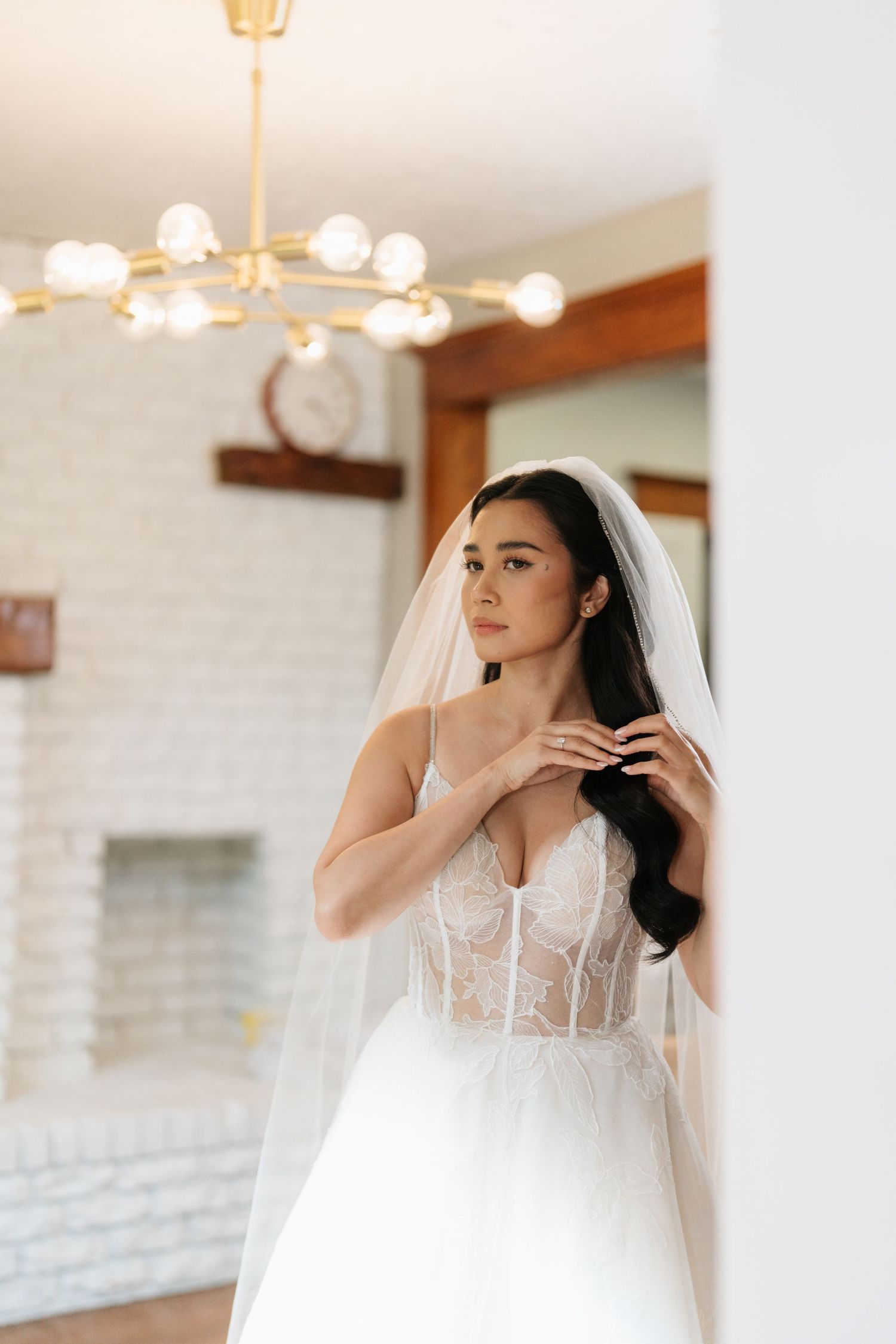 A bride in a white corset wedding dress with sheer details and flowing veil poses in an elegant indoor setting.