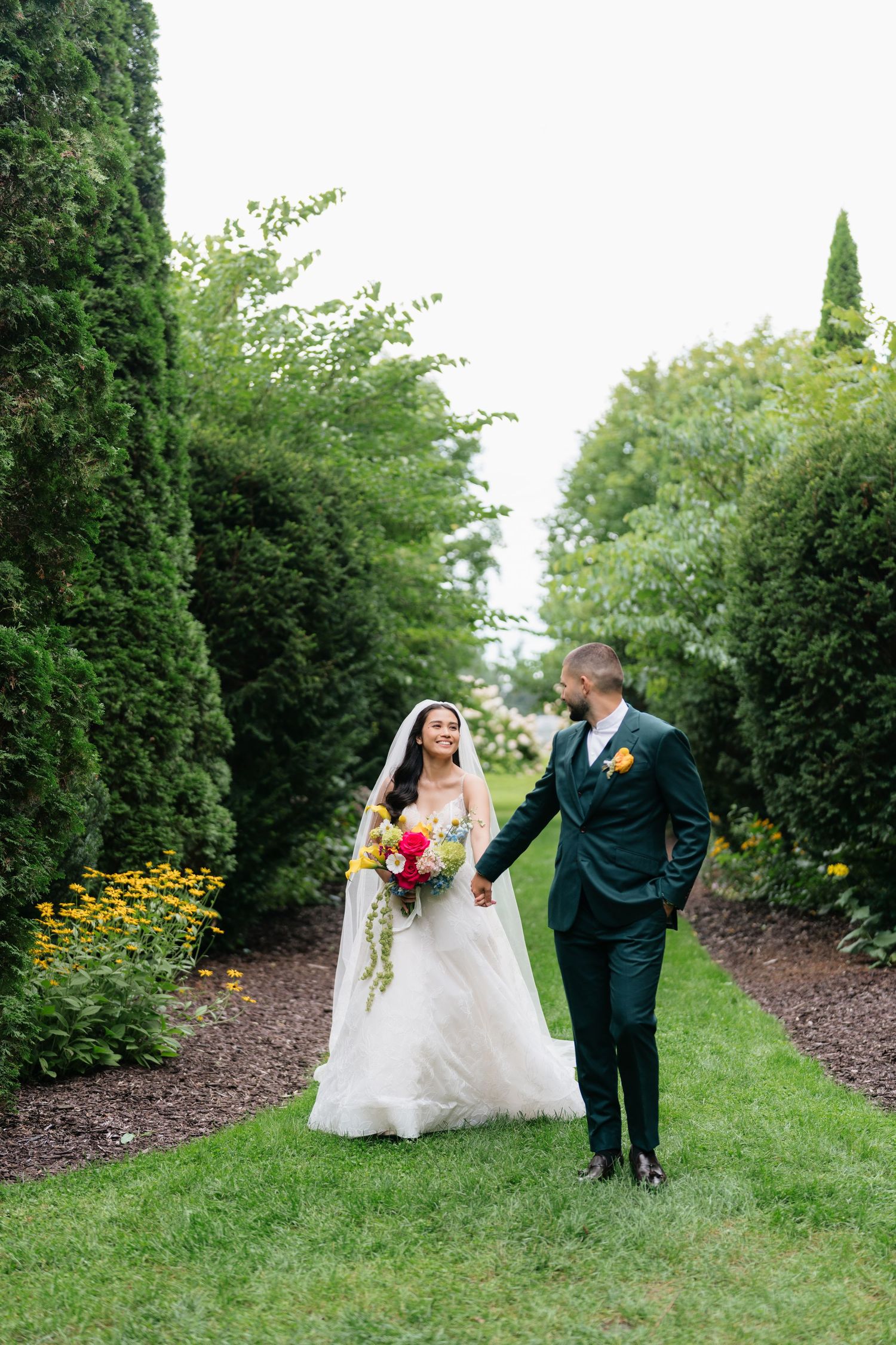 A couple shares romantic moments during their wedding photoshoot in a garden path lined with evergreen trees.