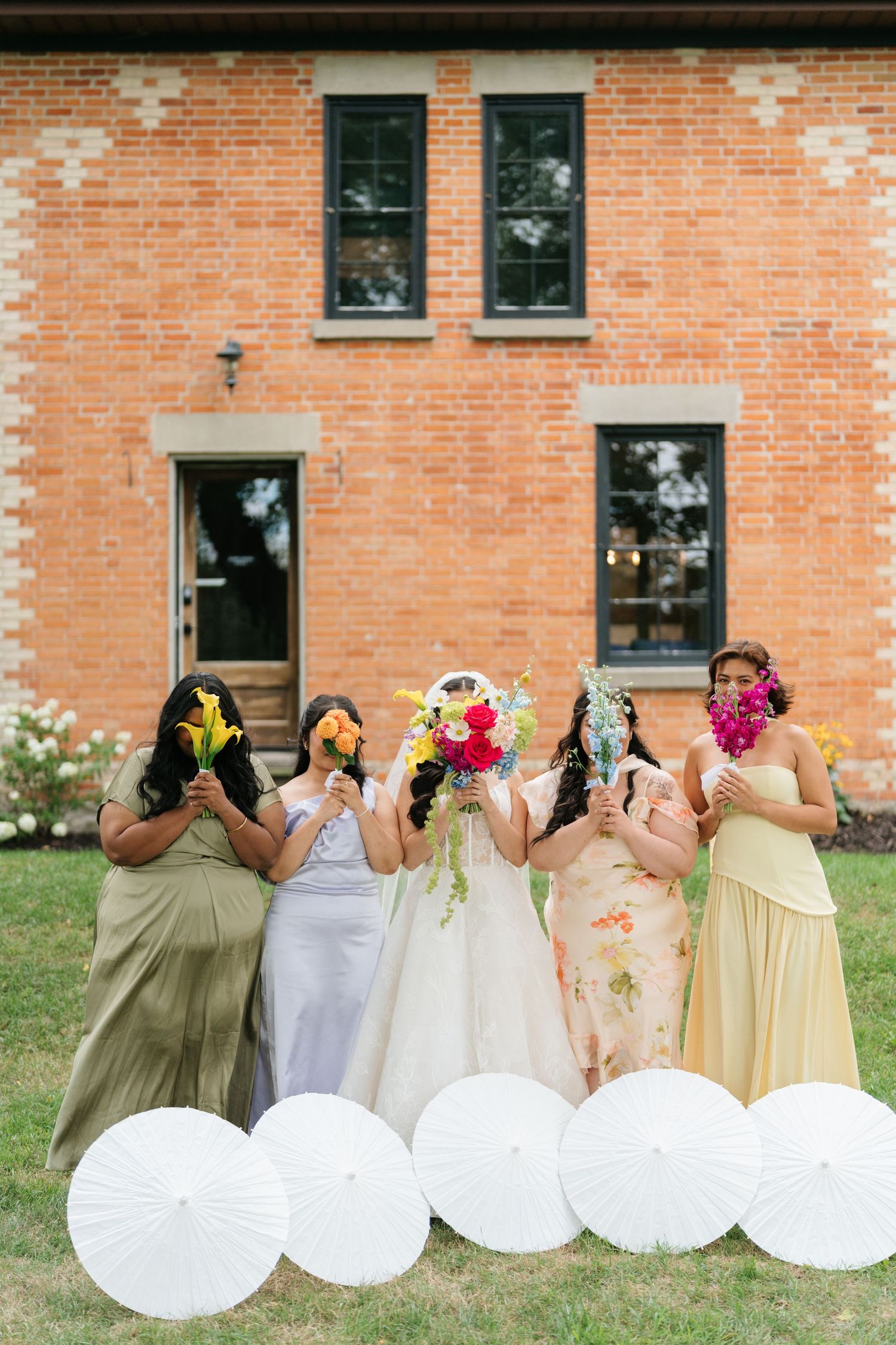 Bridesmaids in yellow and cream dresses sit in a row on grass showing their backs to the camera against brick building.