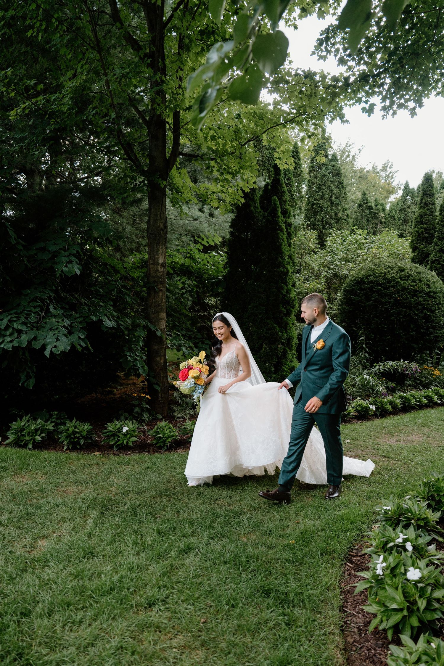 Wedding couple relaxes on grass surrounded by landscaped gardens and tall evergreen trees.