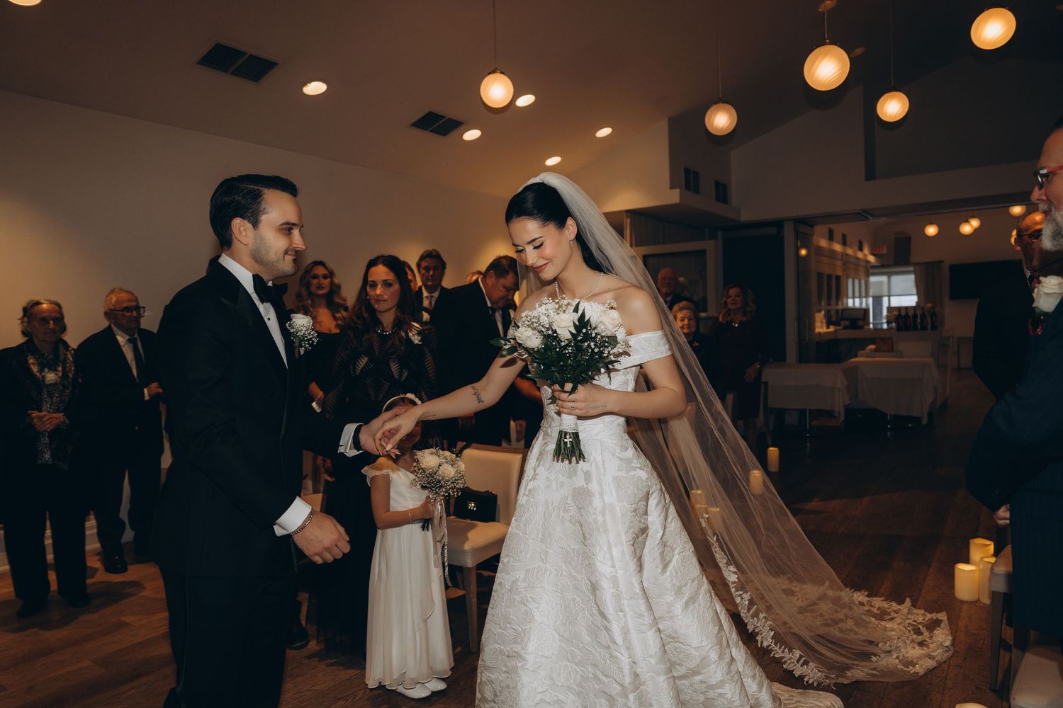 A couple exchanges vows during an intimate indoor wedding ceremony with soft lighting and guests watching in the background.