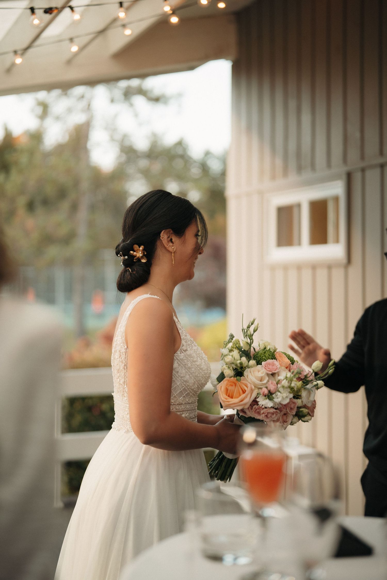 A bride in an elegant white gown holds a bouquet of pink and peach flowers at an outdoor venue.