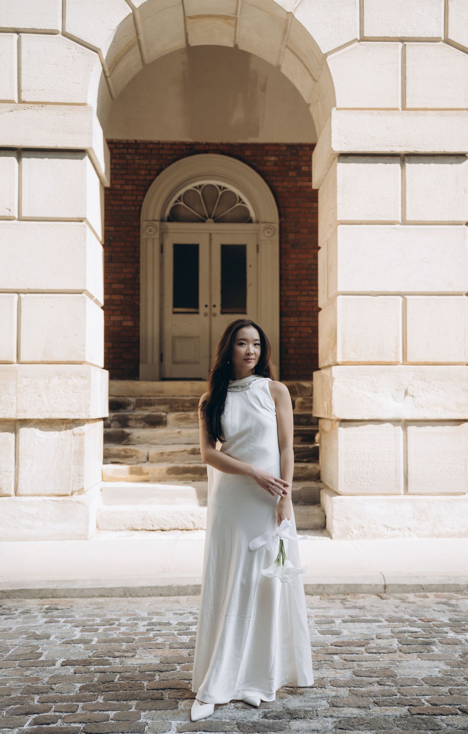 A woman in a flowing white dress poses against an arched brick and stone wall in an elegant architectural setting.