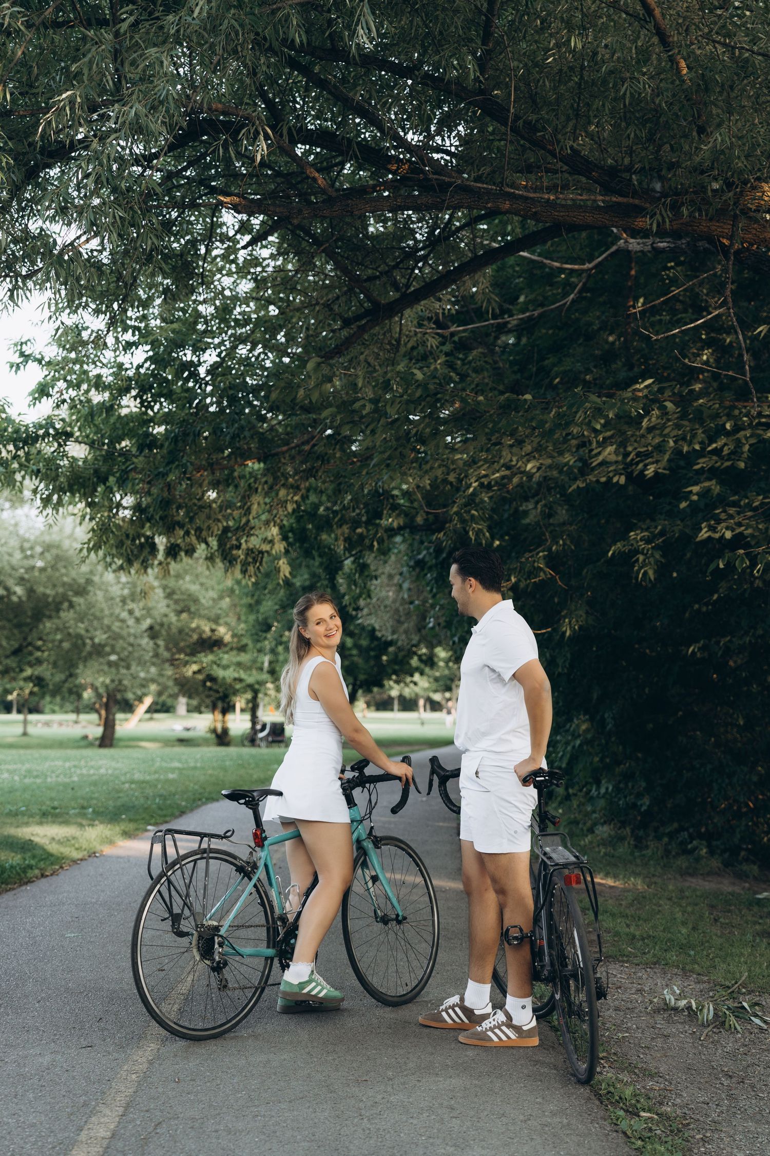 Two people in white summer outfits standing with bicycles on a tree-lined path in a park.