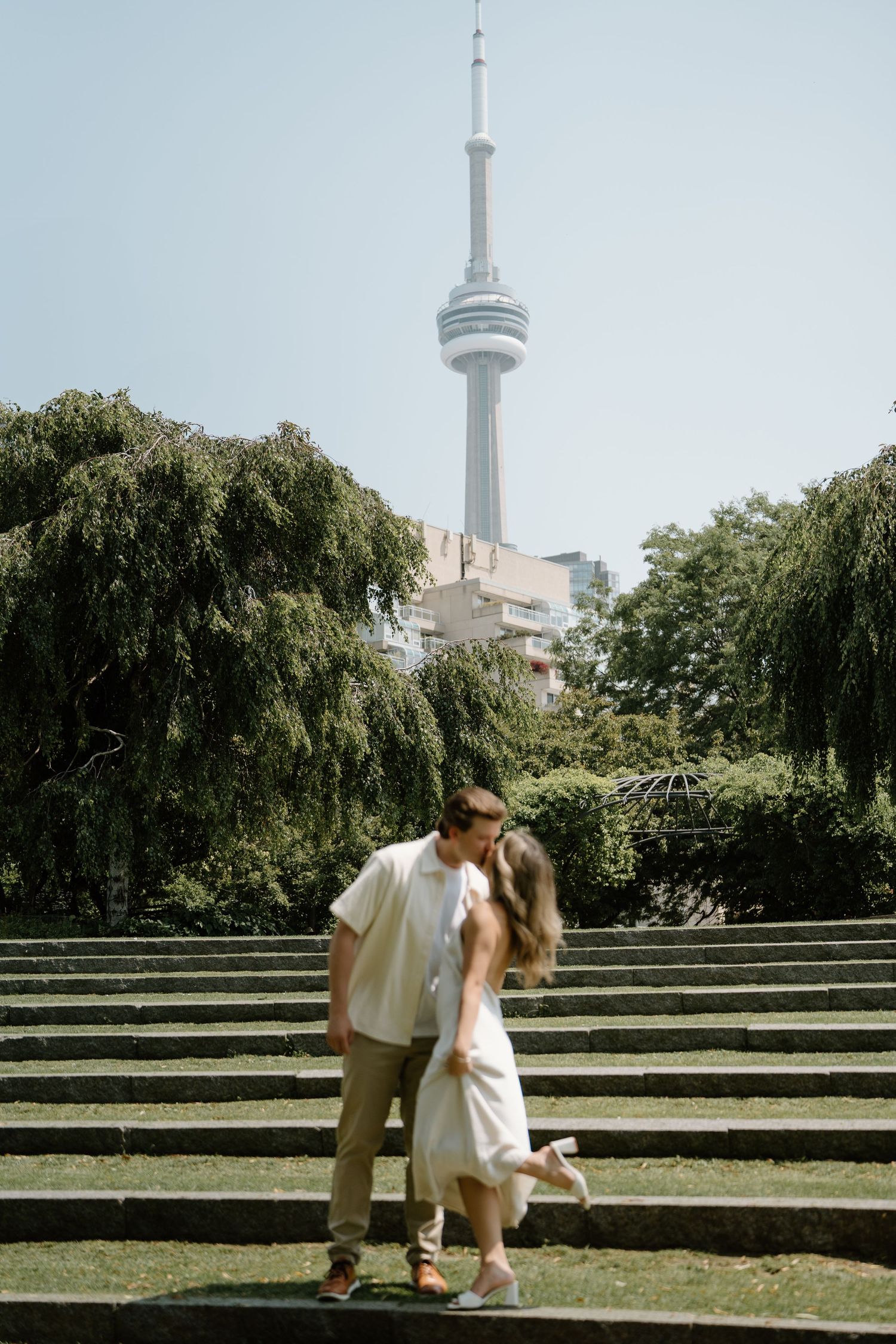 A romantic scene unfolds at a Toronto park with the CN Tower visible in the background beneath weeping willow trees.