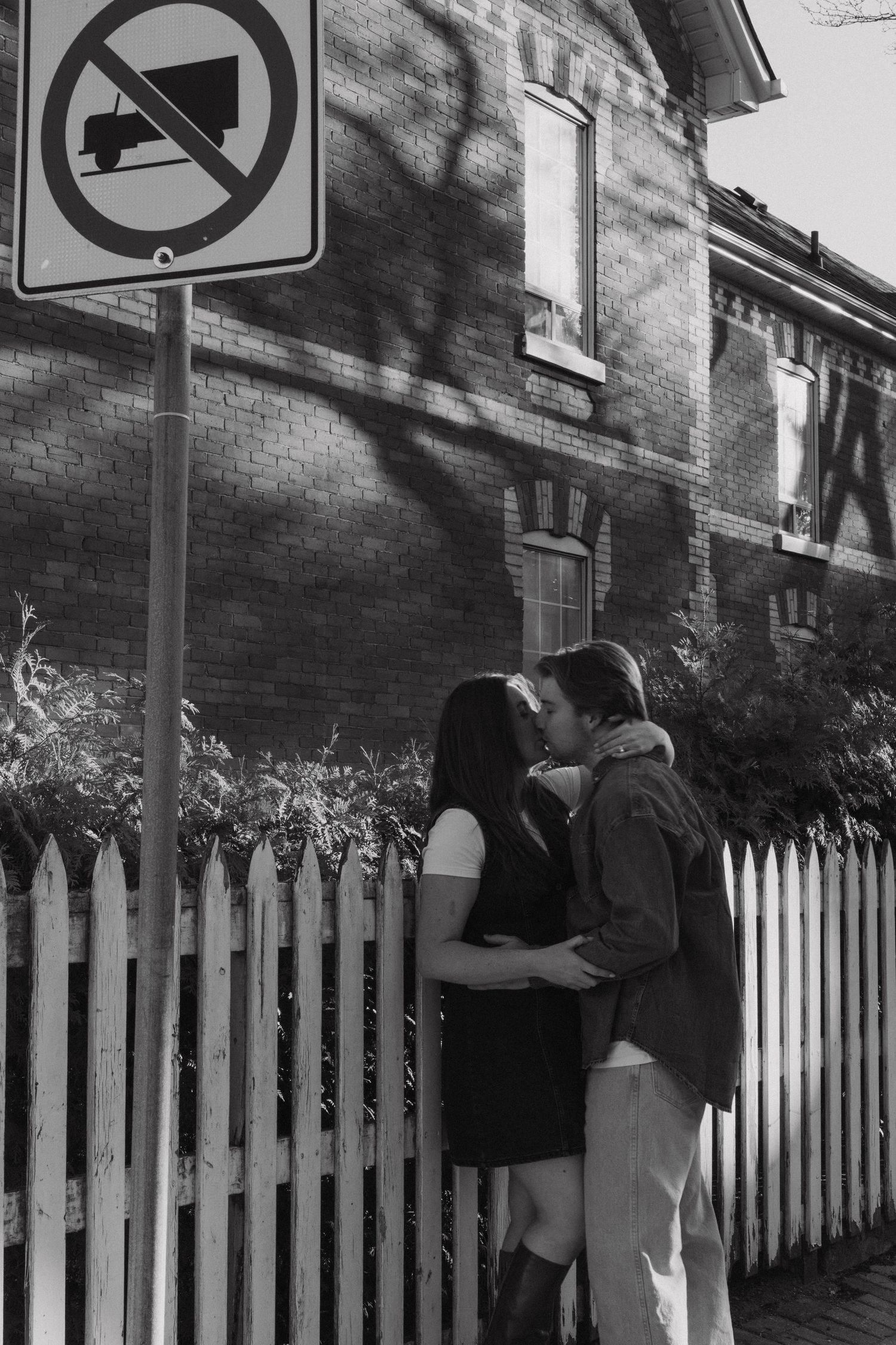 Black and white photo of a romantic embrace by a white picket fence with a no parking sign visible in frame.