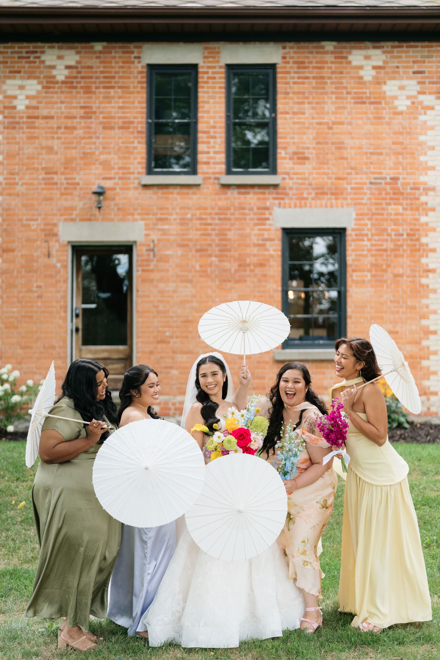 A group in yellow and white dresses holding parasols poses together in front of a brick building.