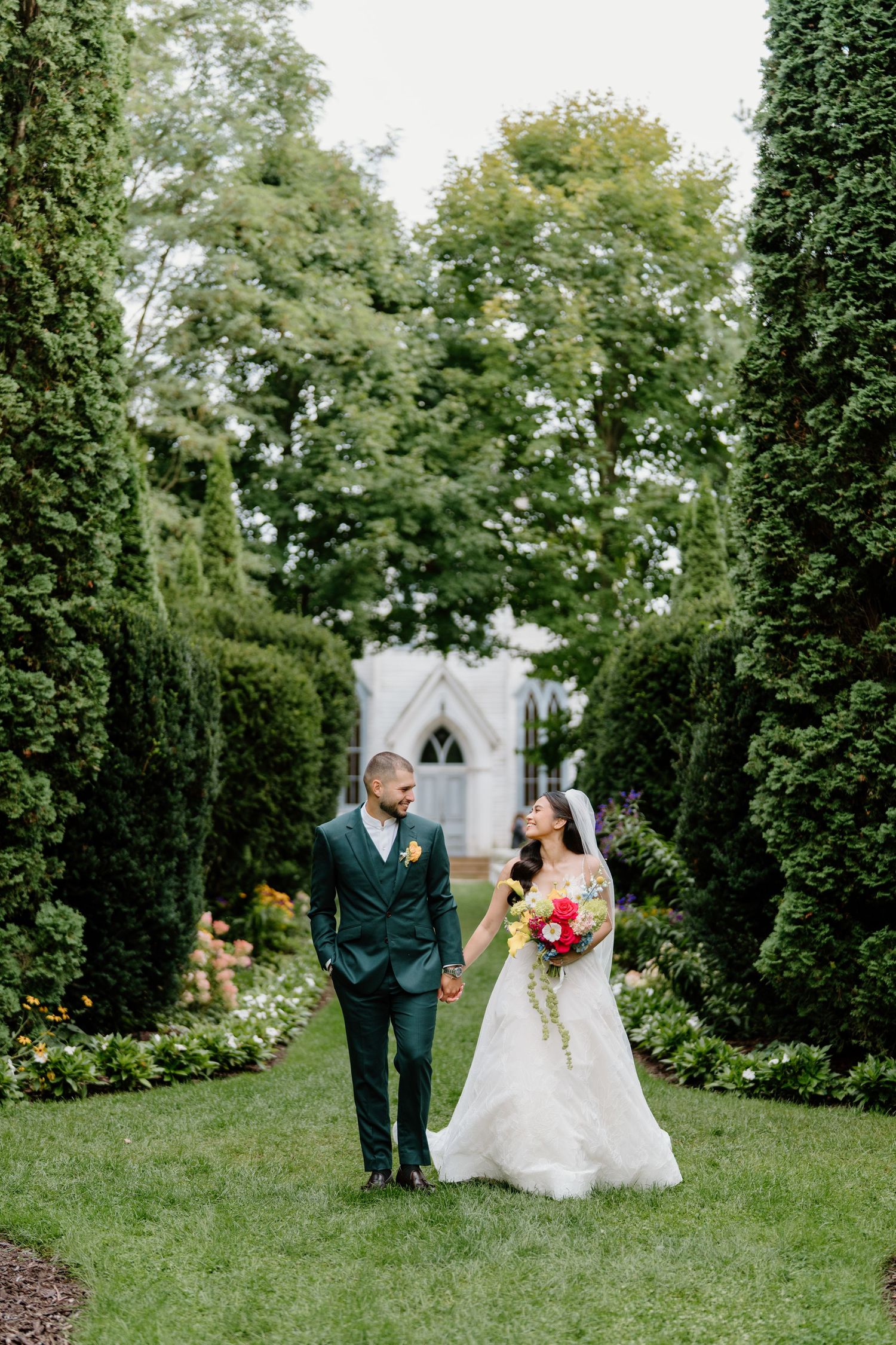 A couple in wedding attire walks through a lush garden pathway surrounded by tall green hedges and trees.