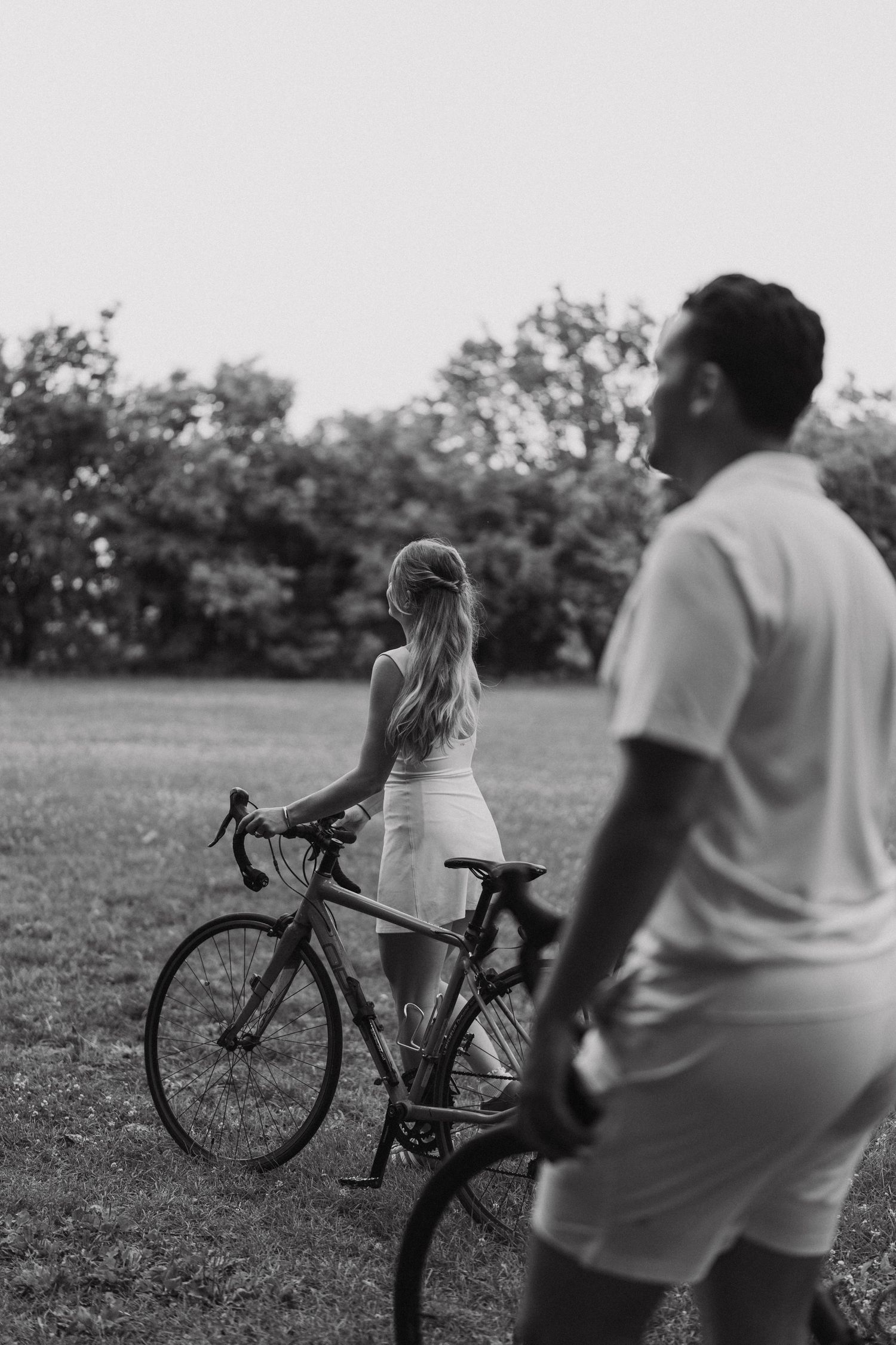 A black and white photo shows two people with bikes standing in a grassy field surrounded by trees.