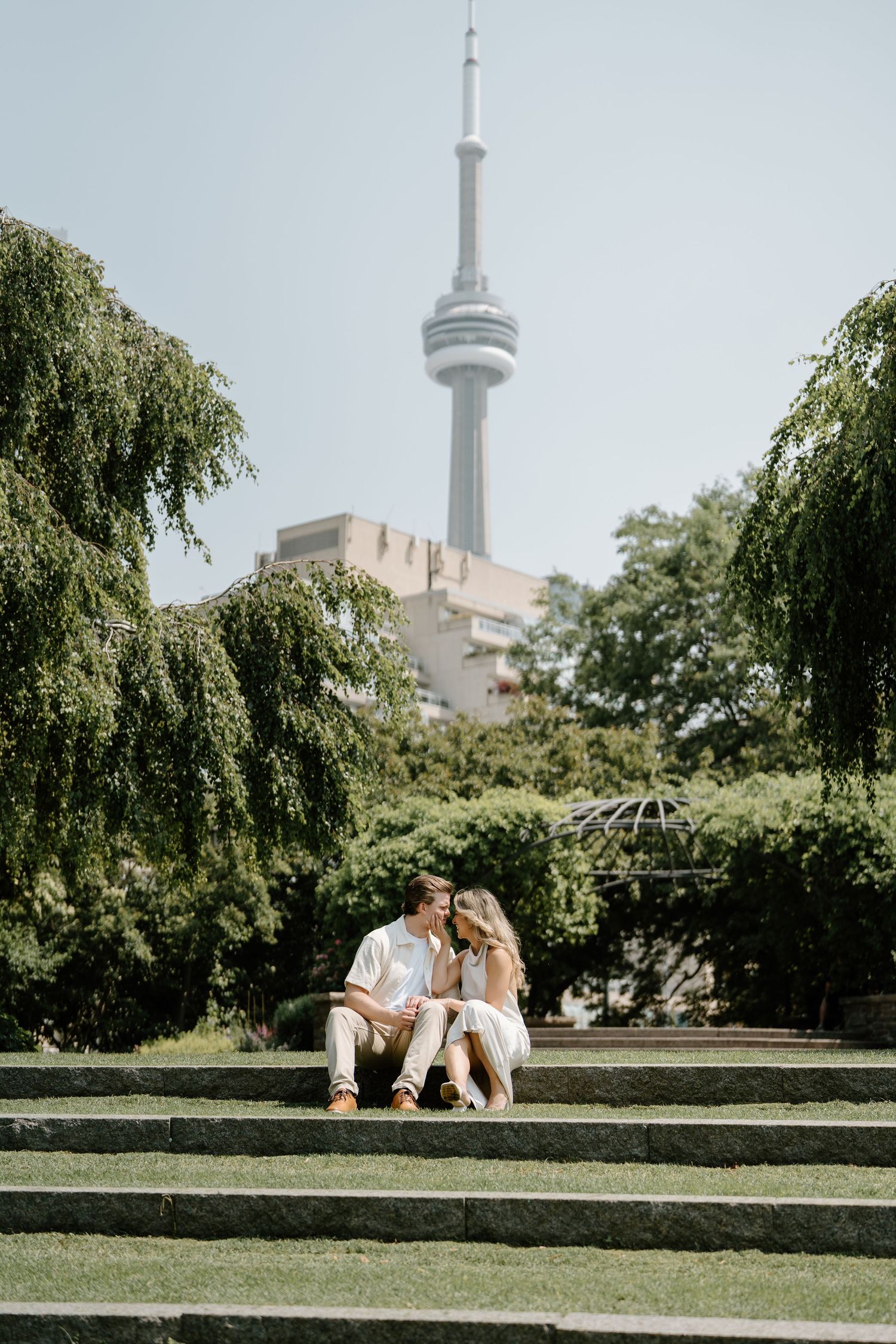 A couple sits on stone steps in a Toronto garden with the CN Tower soaring above the trees.