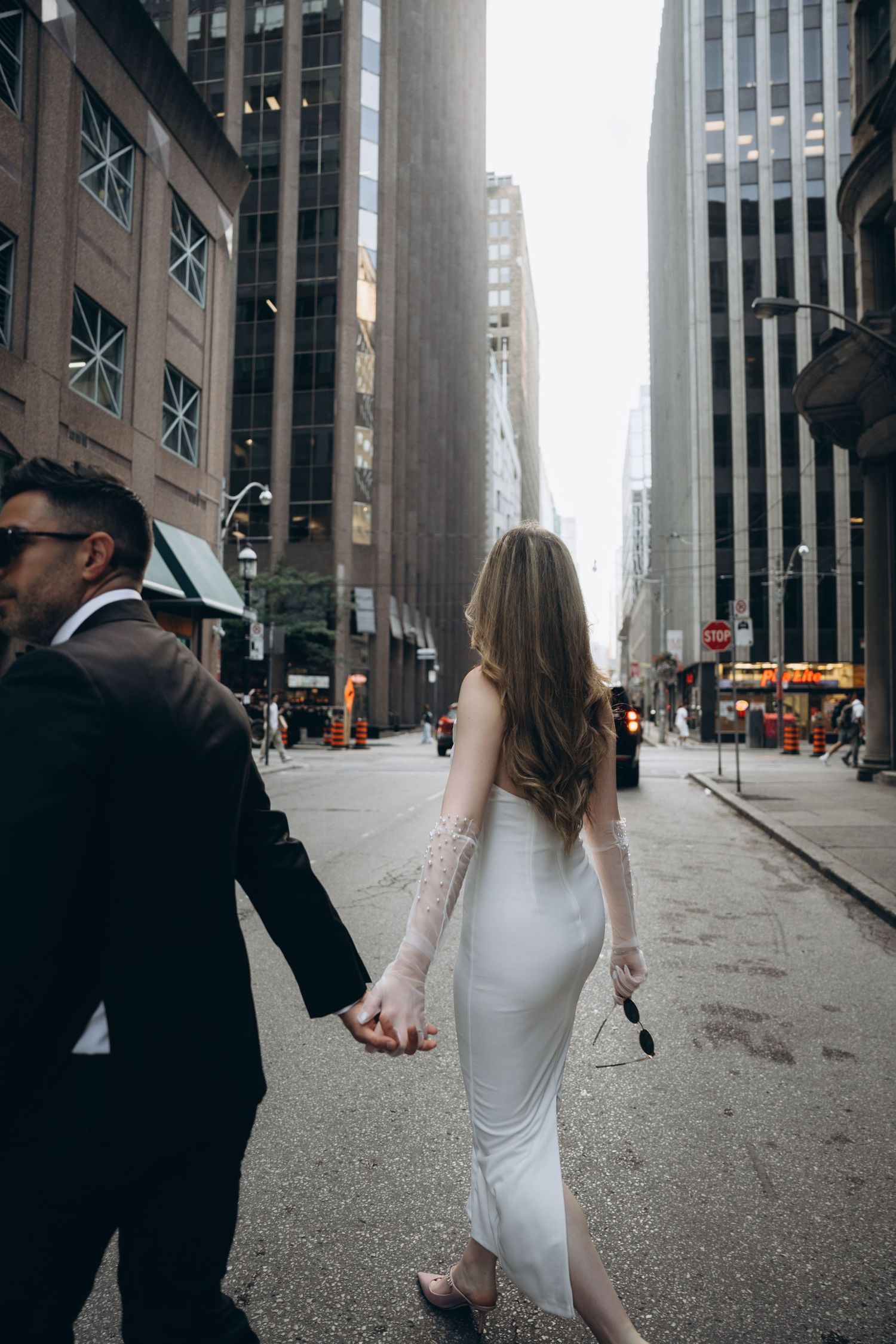 Couple walking on a city street between tall buildings in New York City, holding hands in formal attire.