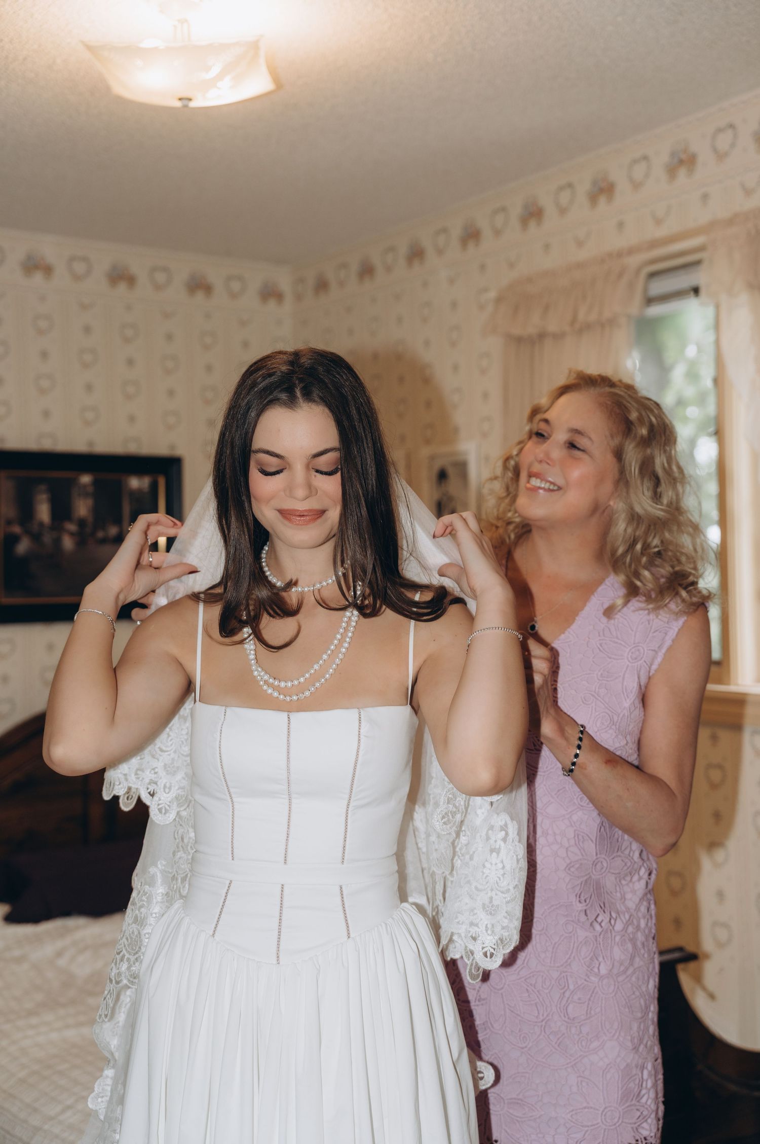 Two people dance and laugh together in a sunlit room with floral wallpaper while wearing formal attire.