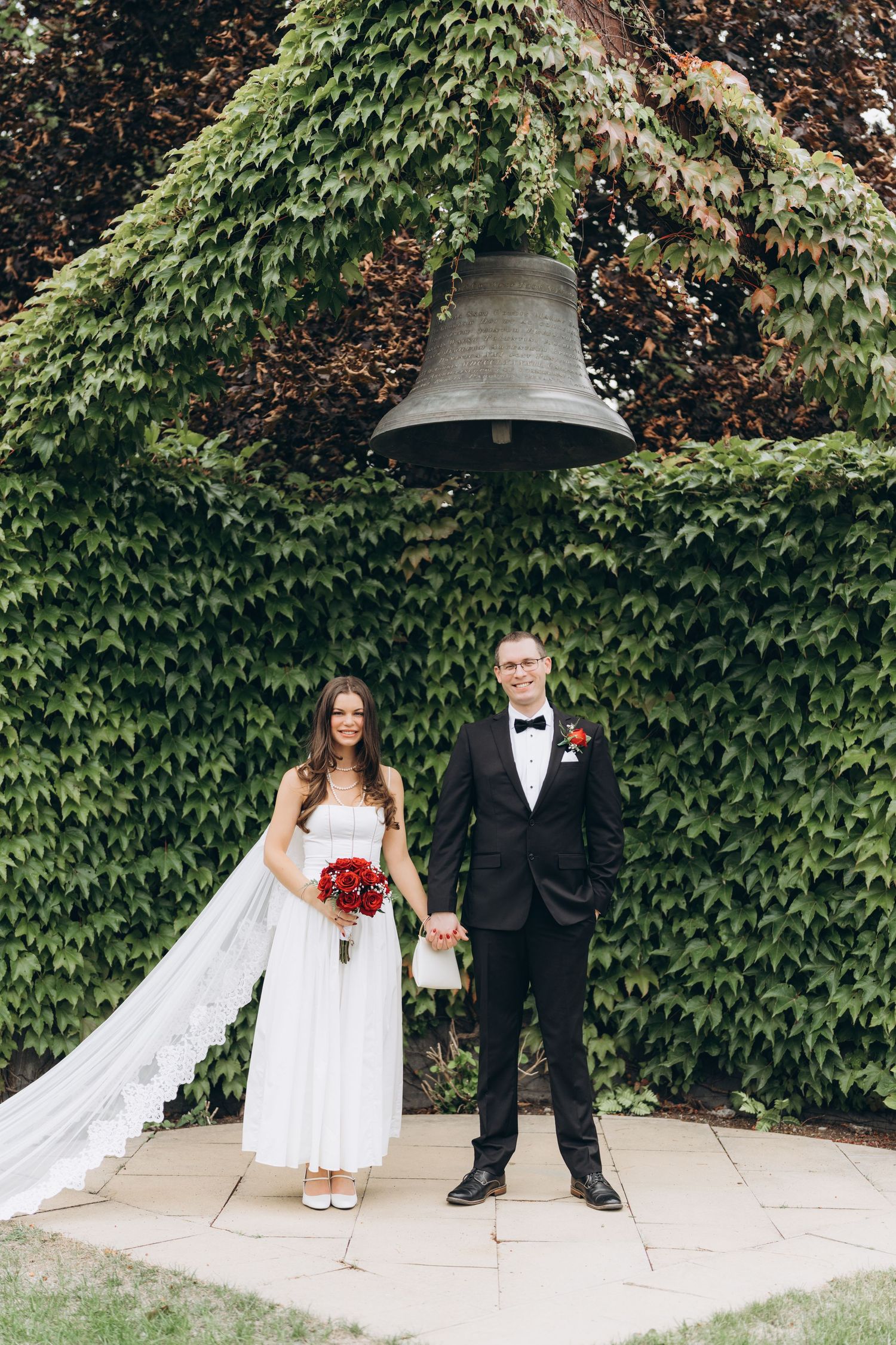 A wedding couple's feet playfully positioned in front of an ivy-covered wall with a church bell.