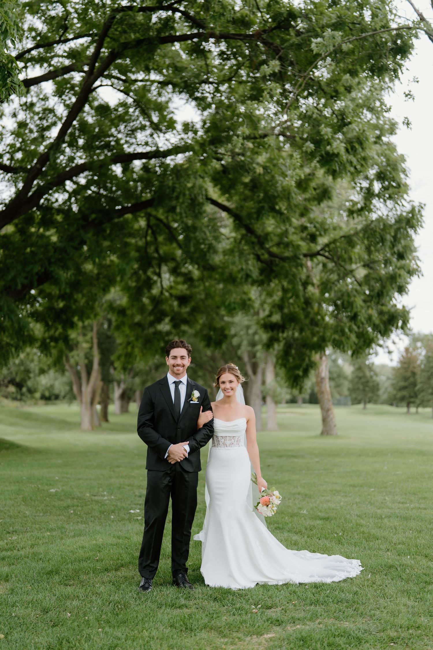 A newlywed couple poses for photos on a grassy hill surrounded by trees.