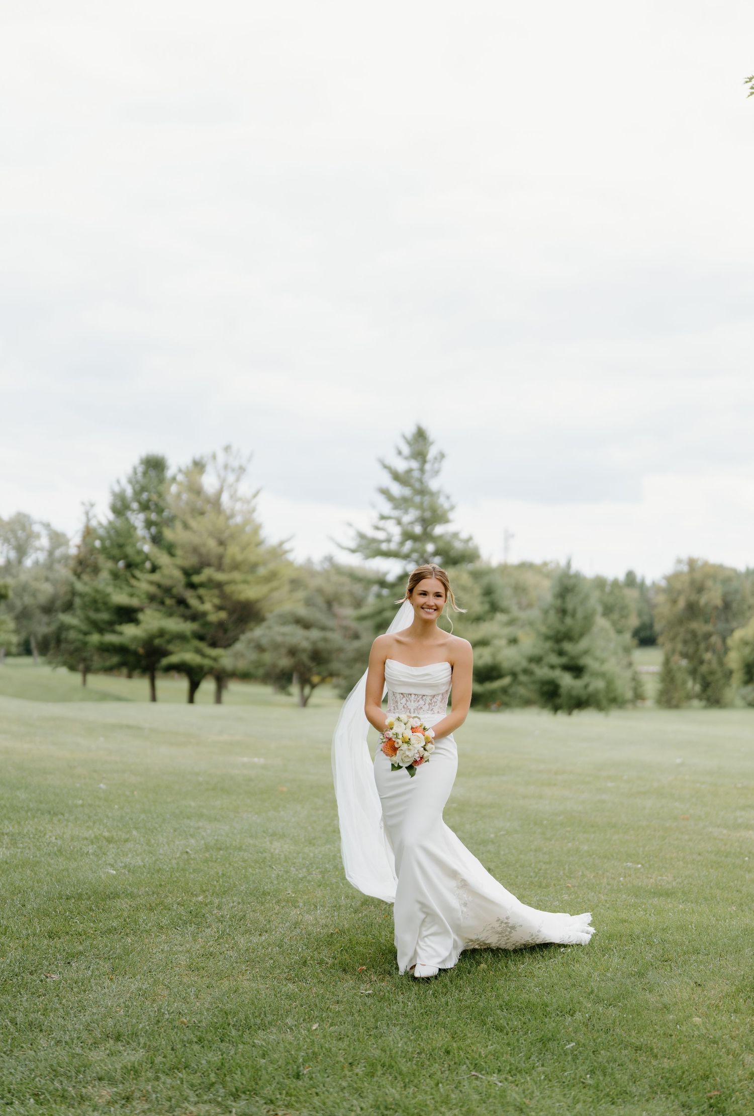 Person in a white wedding dress walking and twirling on a grassy hillside with evergreen trees in the background.
