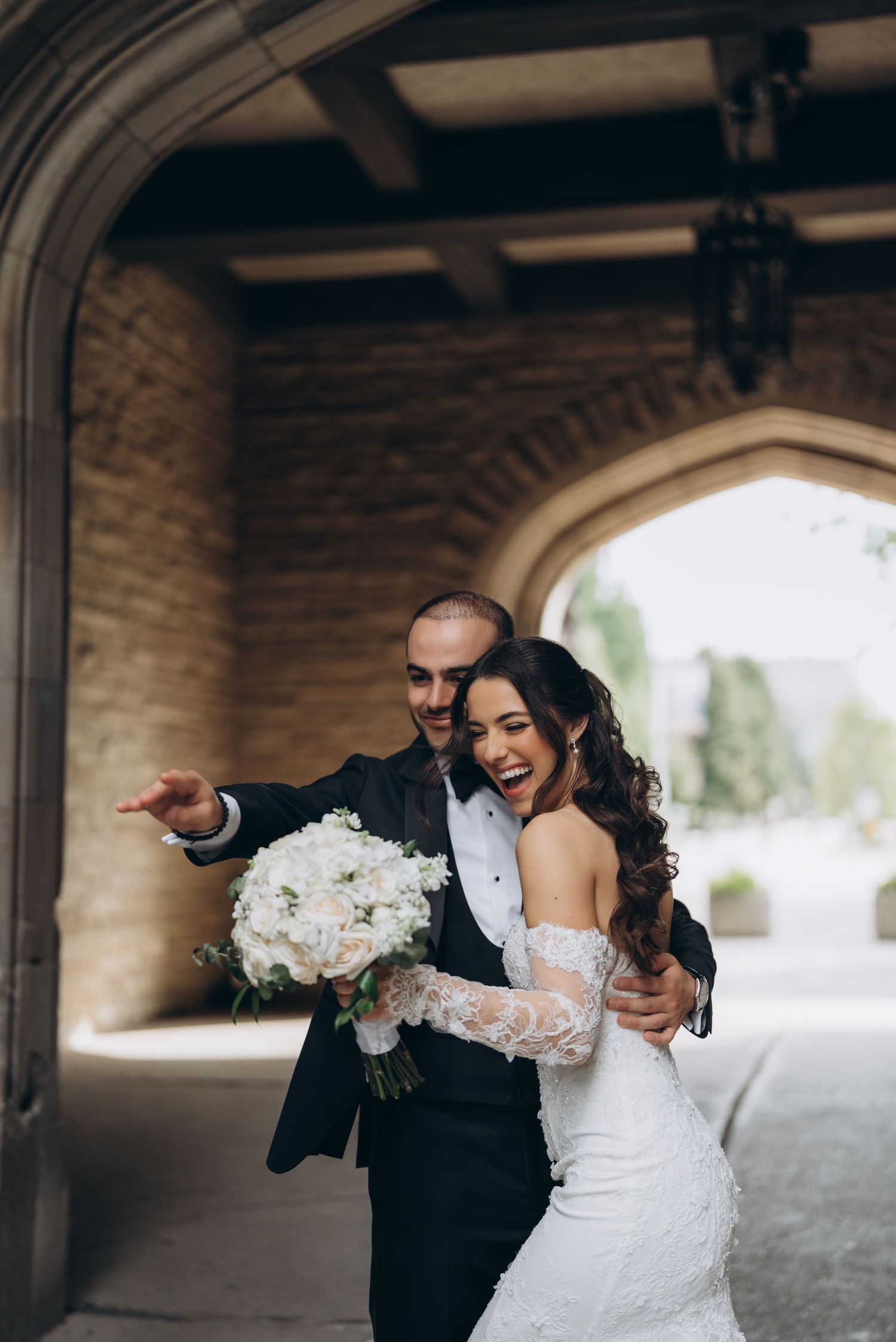 A playful wedding couple shares a candid moment under a covered walkway with white floral bouquets.