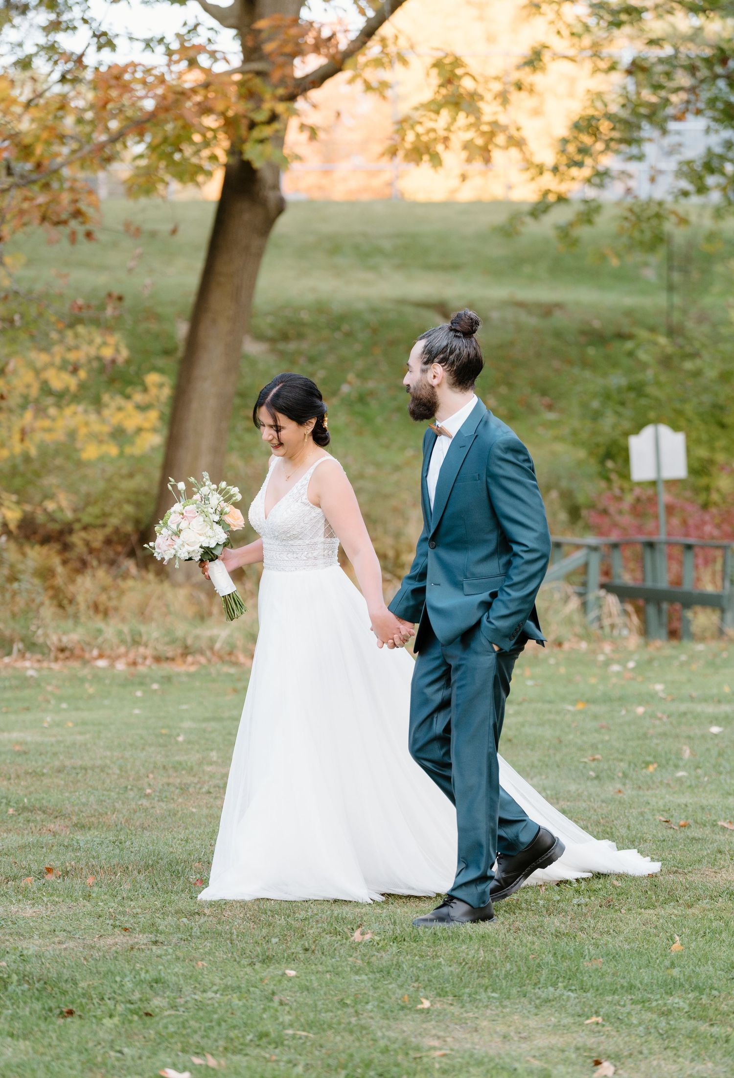 A newlywed couple walks hand in hand across a garden setting in autumn.