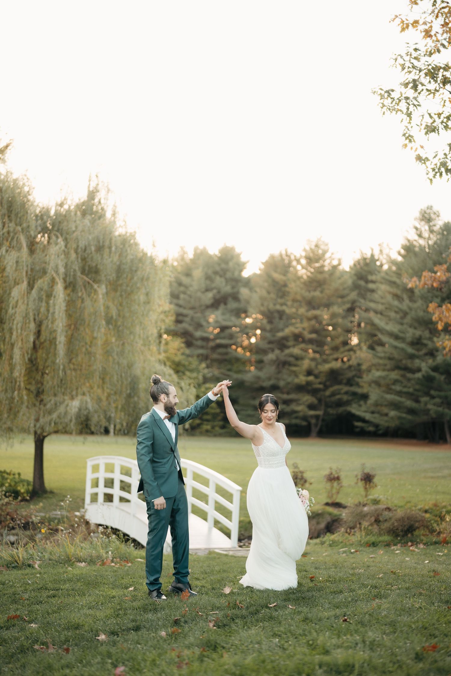 Couple dancing on lawn with white bridge and willow tree in background.