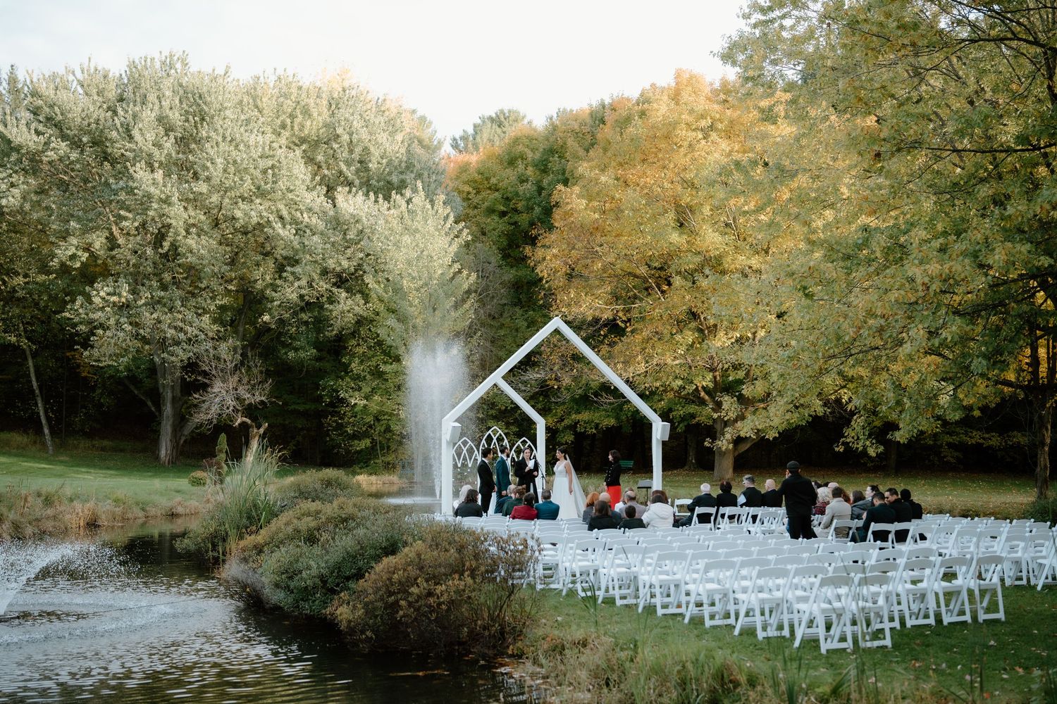 White wedding ceremony setup with chairs near a pond and garden arch against fall foliage.